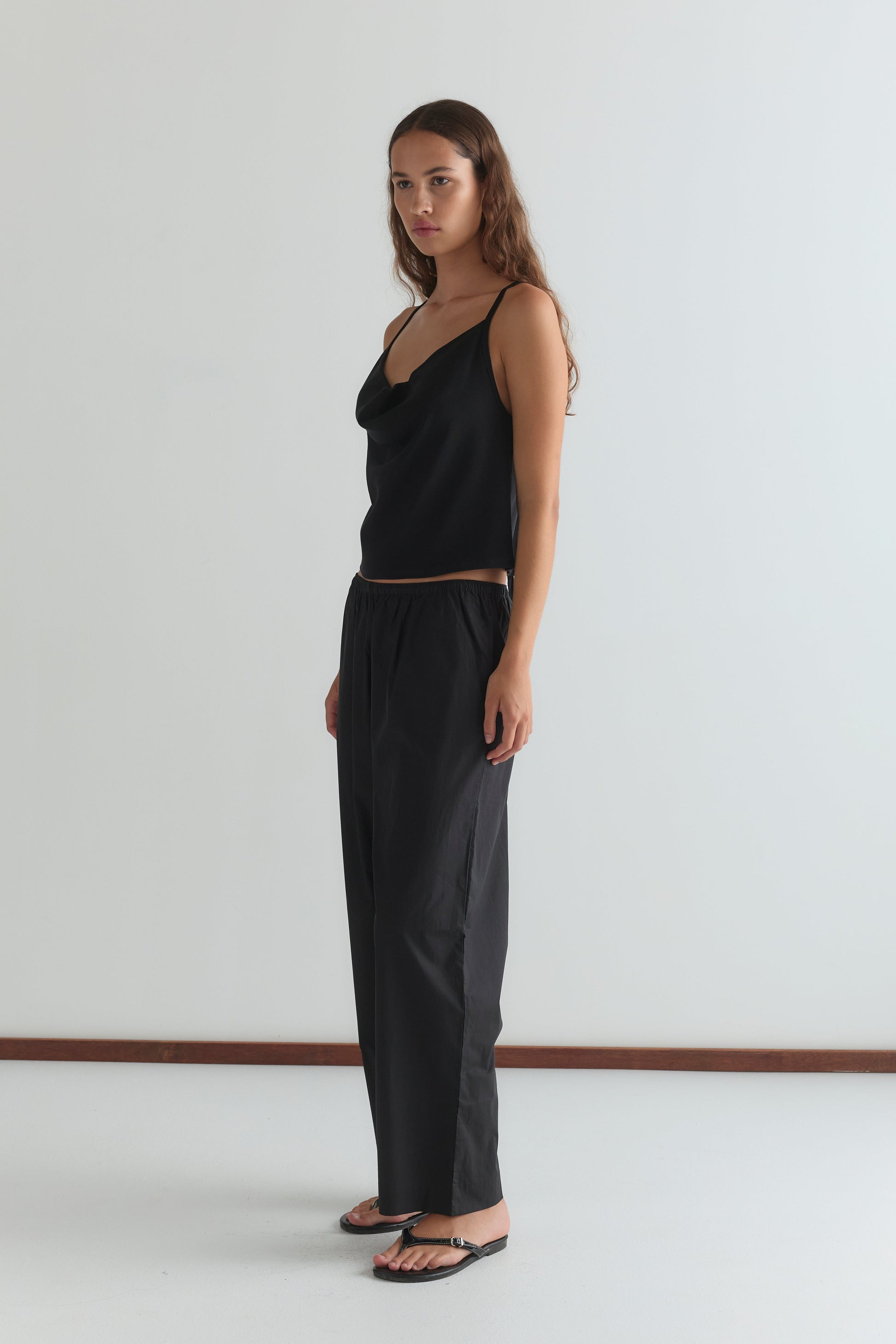 A woman with long brown hair wears the Drape Jersey Top - Black by Deiji Studios, paired with black pants and sandals, standing against a plain white wall and looking slightly to the side with a neutral expression.