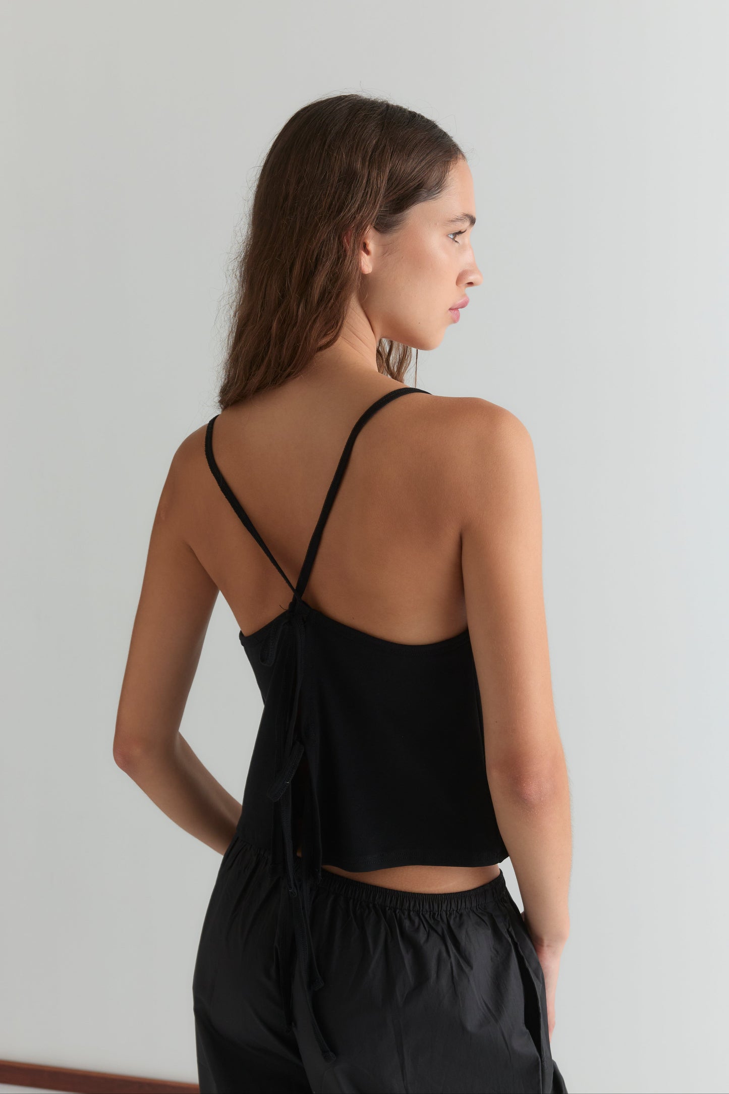 A woman with wavy brown hair stands sideways, facing away, wearing a black Drape Jersey Top by Deiji Studios with an open back and black pants against a plain light background.
