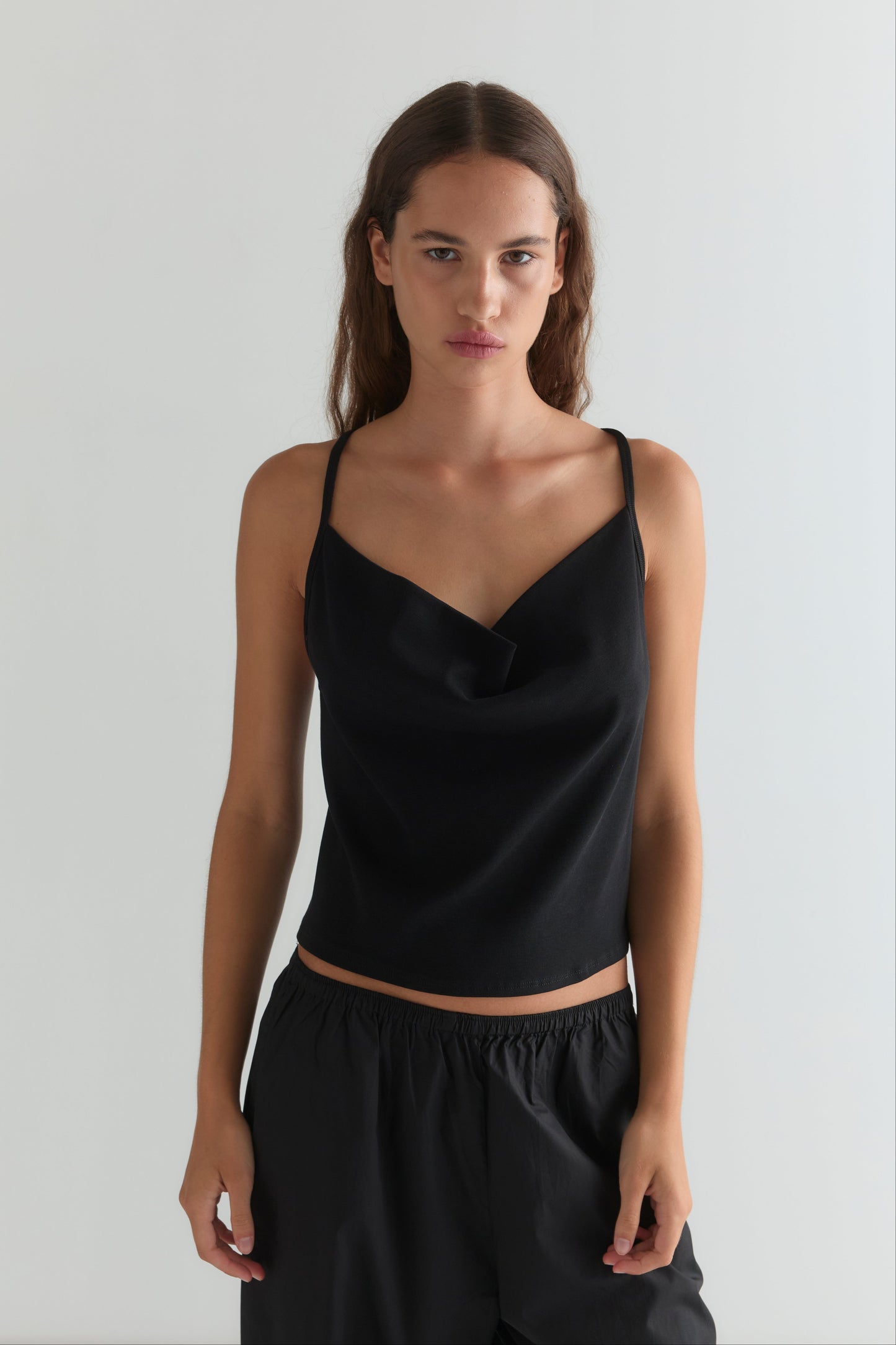 A woman with long brown hair and a neutral expression wears the Drape Jersey Top - Black by Deiji Studios, featuring thin straps, paired with loose black pants, standing against a plain white background.
