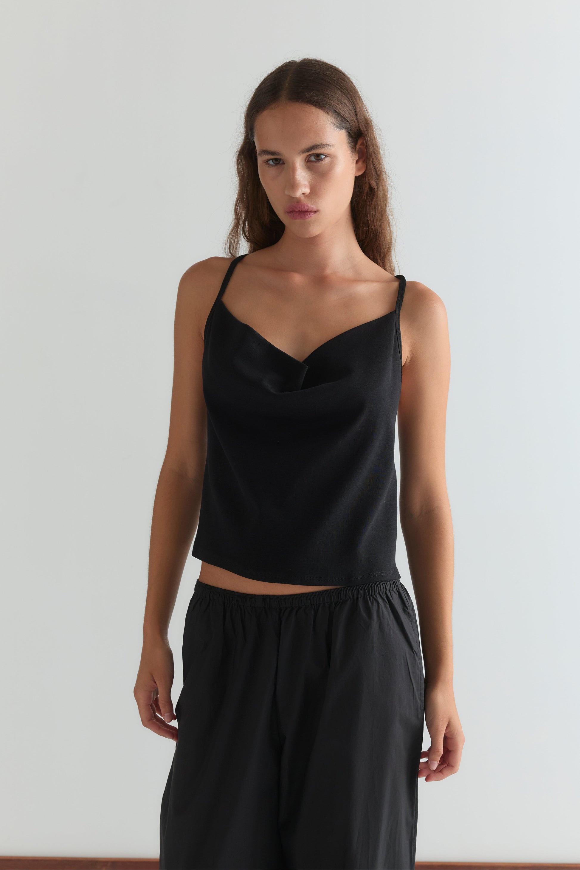 A woman with long brown hair wears the Drape Jersey Top - Black by Deiji Studios and loose black pants, standing against a plain white background and looking directly at the camera with a neutral expression.