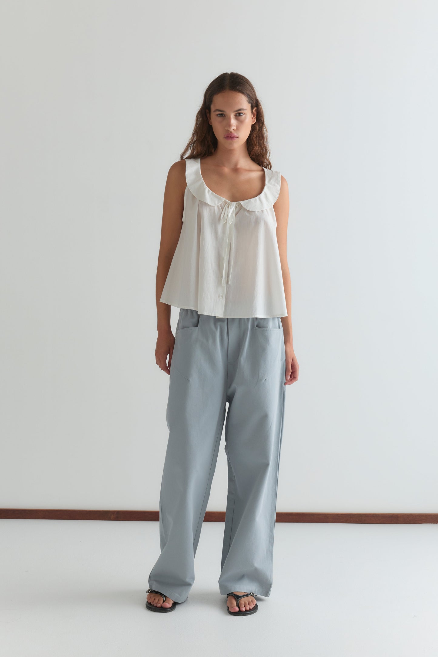 A woman with long brown hair stands against a white wall, wearing the Round Collar Top - White Texture by Deiji Studios, styled with light blue loose-fitting pants and black sandals.