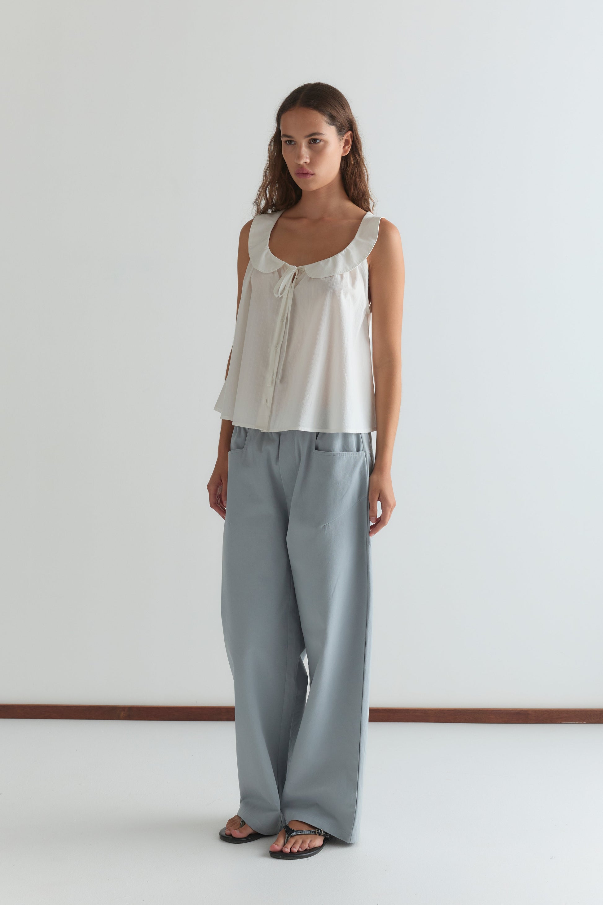 A woman with long brown hair and a neutral expression stands against a white wall wearing the Round Collar Top - White Texture, featuring a ruffled collar and tie detail, paired with light blue wide-leg pants and black sandals.