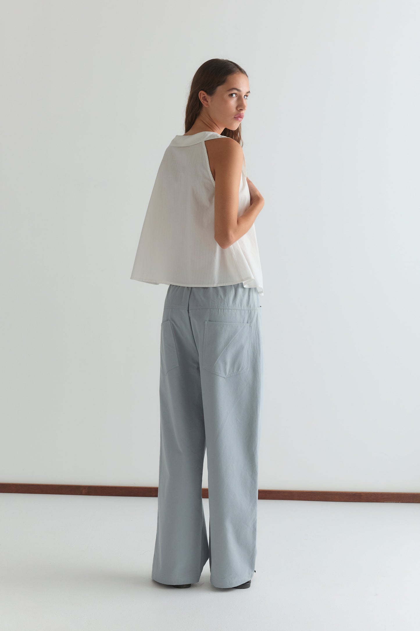 A woman stands facing away, looking over her shoulder. She wears the Deiji Studios Round Collar Top - White Texture and wide-leg light blue pants. The minimal background features white walls and a brown baseboard.