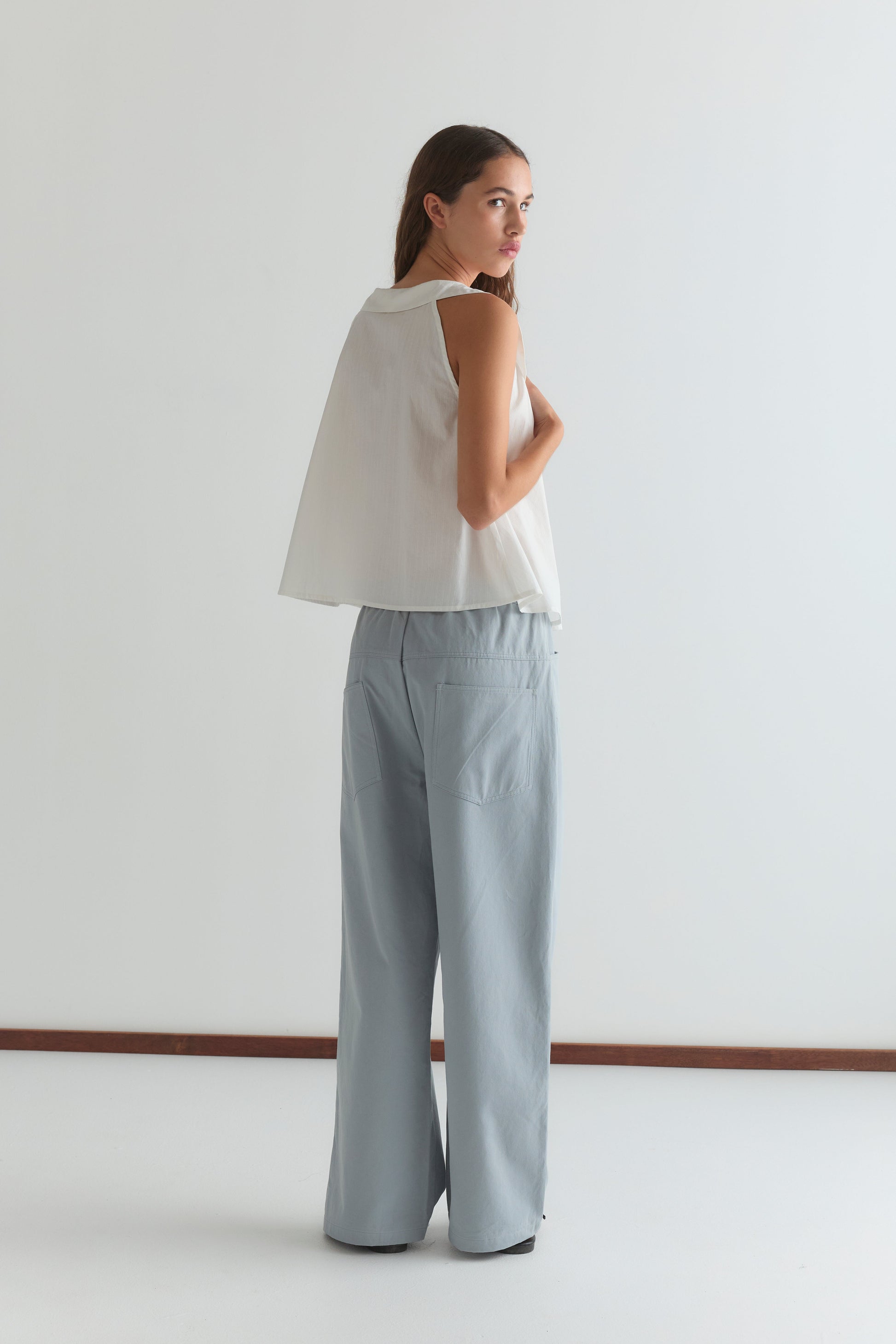 A woman stands facing away, looking over her shoulder. She wears the Deiji Studios Round Collar Top - White Texture and wide-leg light blue pants. The minimal background features white walls and a brown baseboard.
