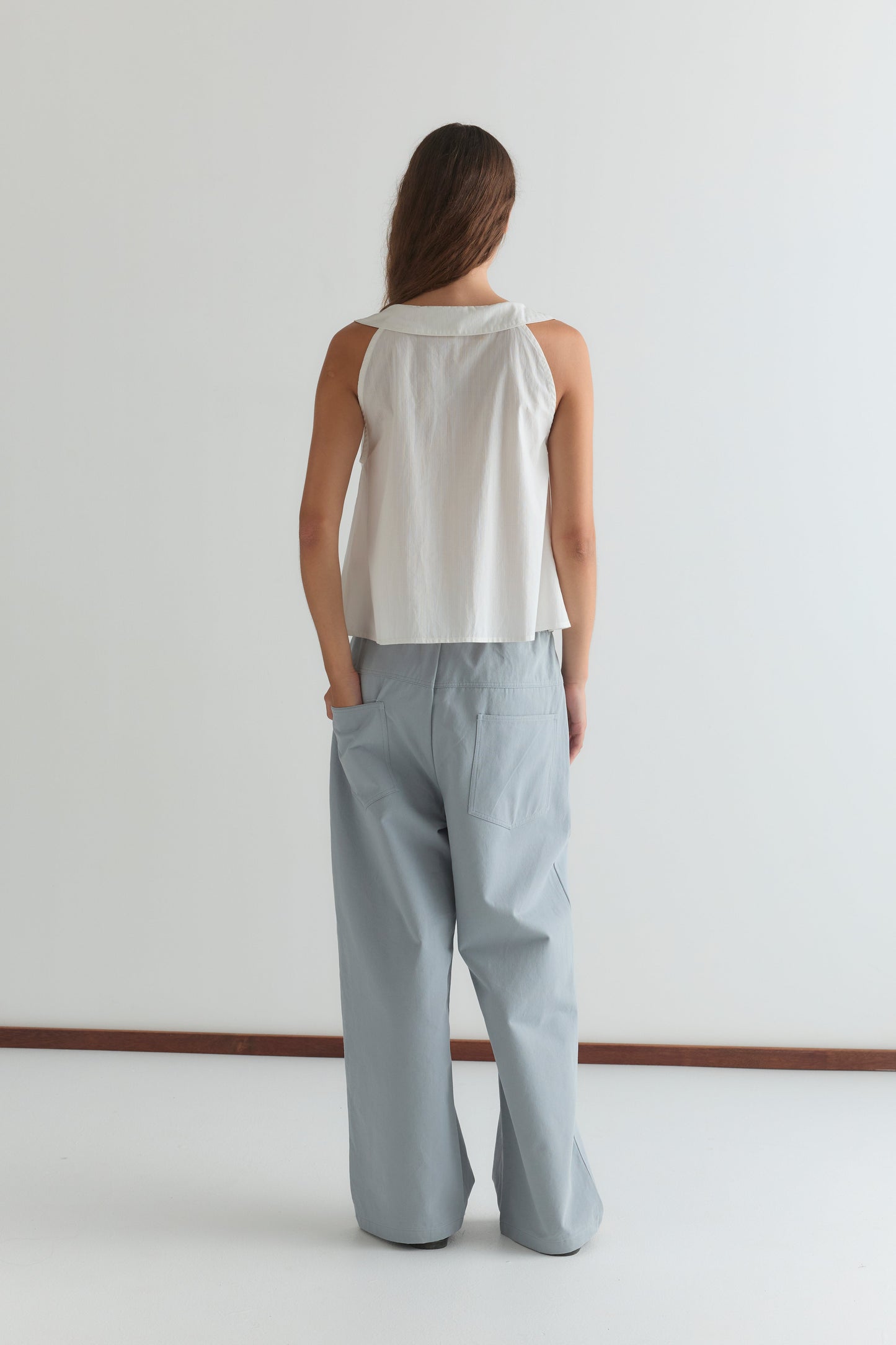 A person with long hair stands facing away, wearing Deiji Studios' Round Collar Top - White Texture and loose light blue pants, hands in pockets, against a plain white background.