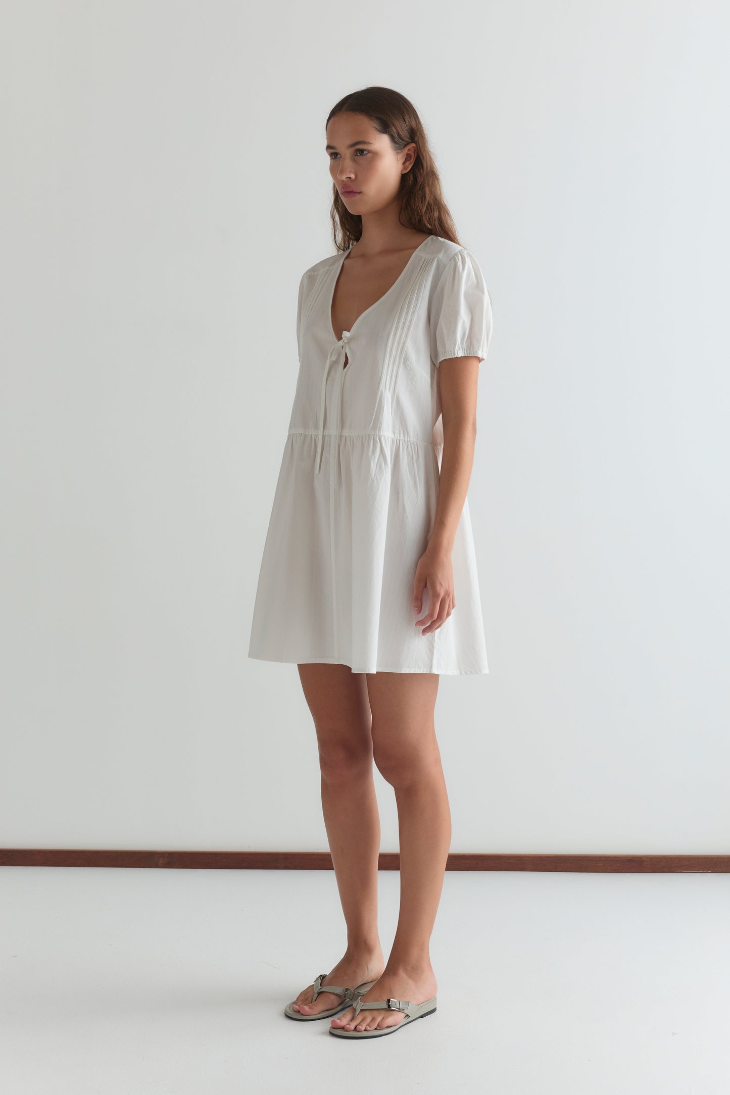 A woman stands indoors against a white wall, wearing the V Pleat Dress - White Texture—a short mini dress with a V-neckline, short sleeves, and a neck tie—paired with flat sandals.
