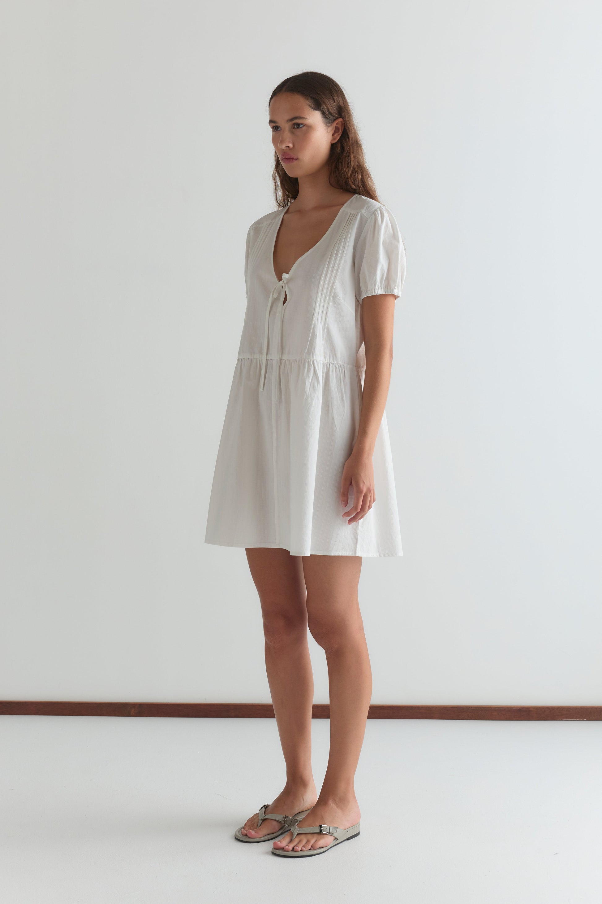 A woman stands indoors against a white wall, wearing the V Pleat Dress - White Texture—a short mini dress with a V-neckline, short sleeves, and a neck tie—paired with flat sandals.