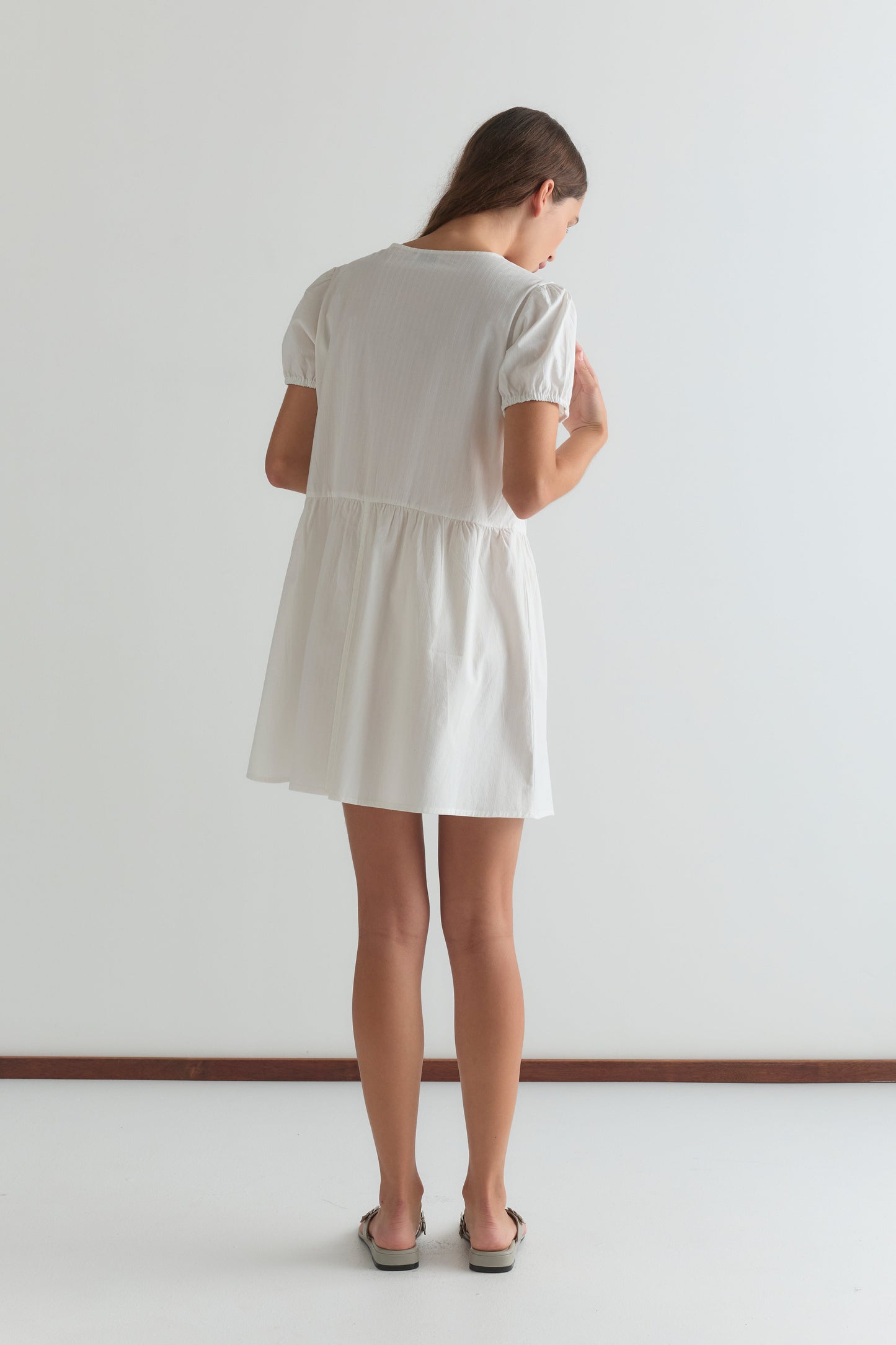 A woman with long brown hair faces away, wearing the V Pleat Dress - White Texture from Deiji Studios with short sleeves and flat sandals, set against a plain white background.