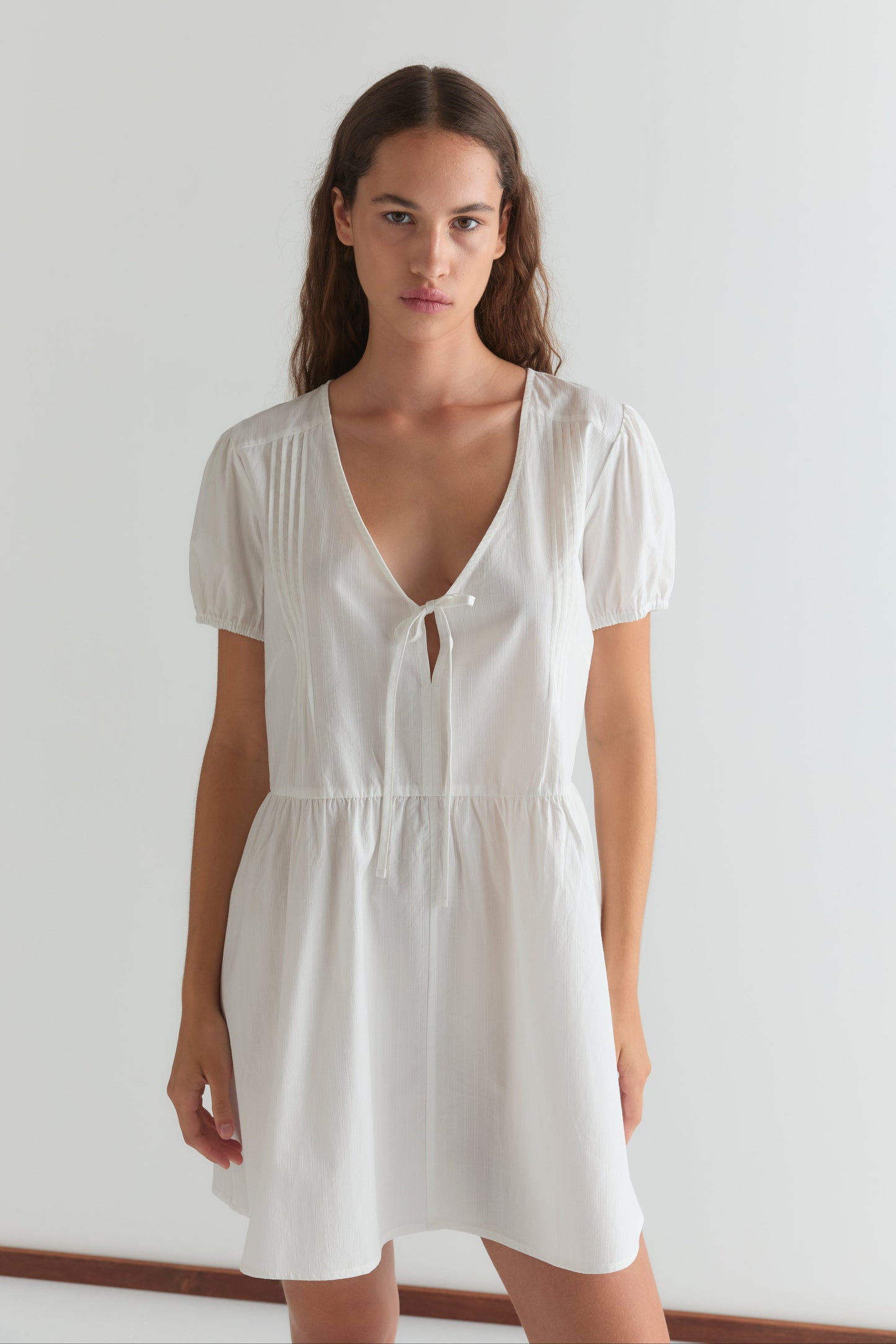 A woman with long brown hair wears the V Pleat Dress - White Texture by Deiji Studios, featuring short puff sleeves and a V-neckline with a small front tie. She stands against a plain white background, looking directly at the camera.