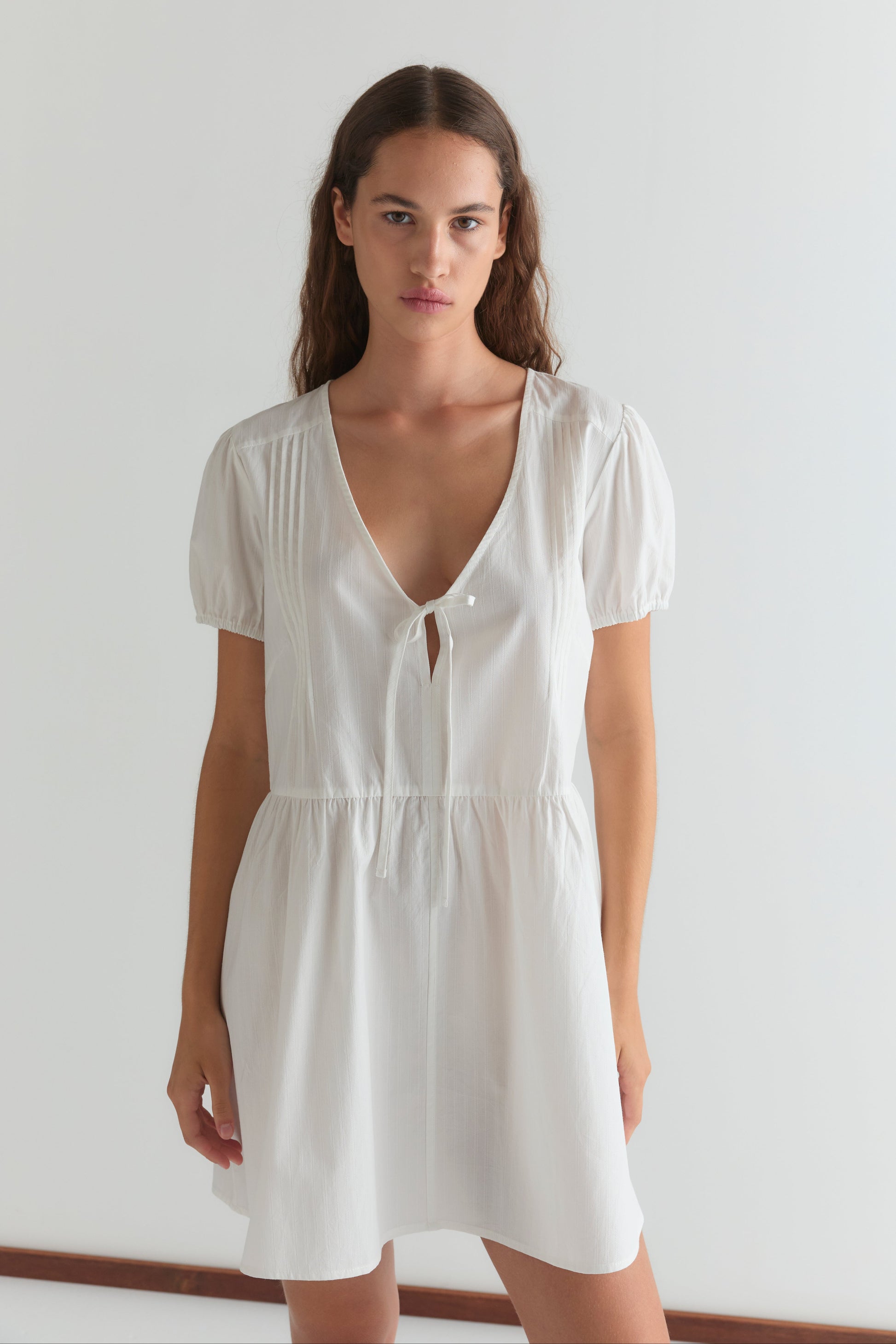 A woman with long brown hair wears the V Pleat Dress - White Texture by Deiji Studios, featuring short puff sleeves and a V-neckline with a small front tie. She stands against a plain white background, looking directly at the camera.