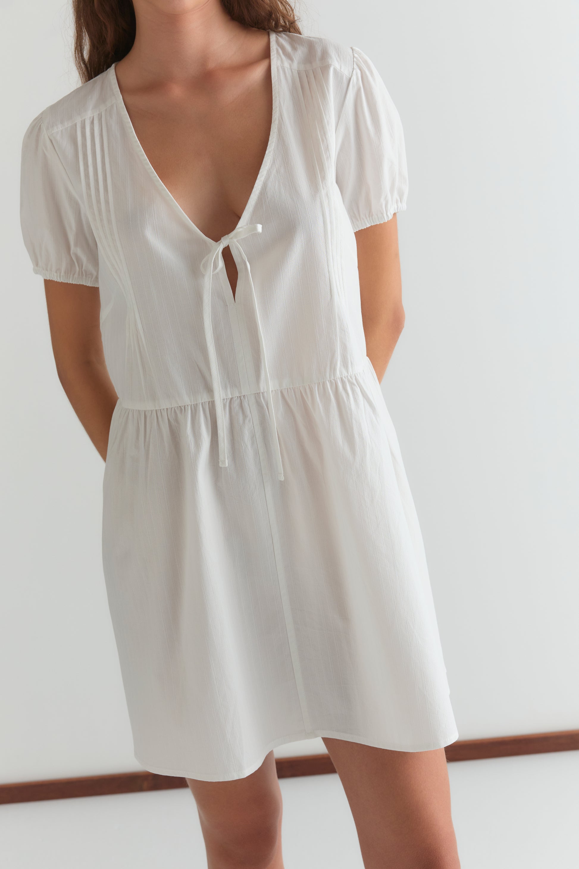 Someone wearing the V Pleat Dress - White Texture from Deiji Studios, a short-sleeved white dress with a deep V-neck, tie detail at the front, and pleats, stands against a plain light background.