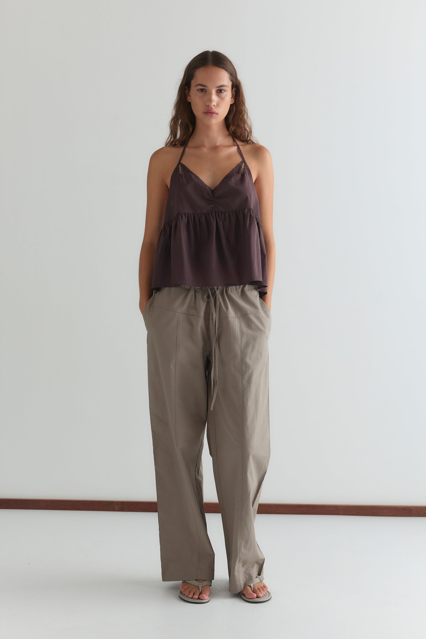 A woman with long, wavy brown hair stands against a white background, wearing the Keyhole Halter Top - Wine by Deiji Studios with adjustable straps, loose beige pants, and sandals. She has a neutral expression and hands in her pockets.