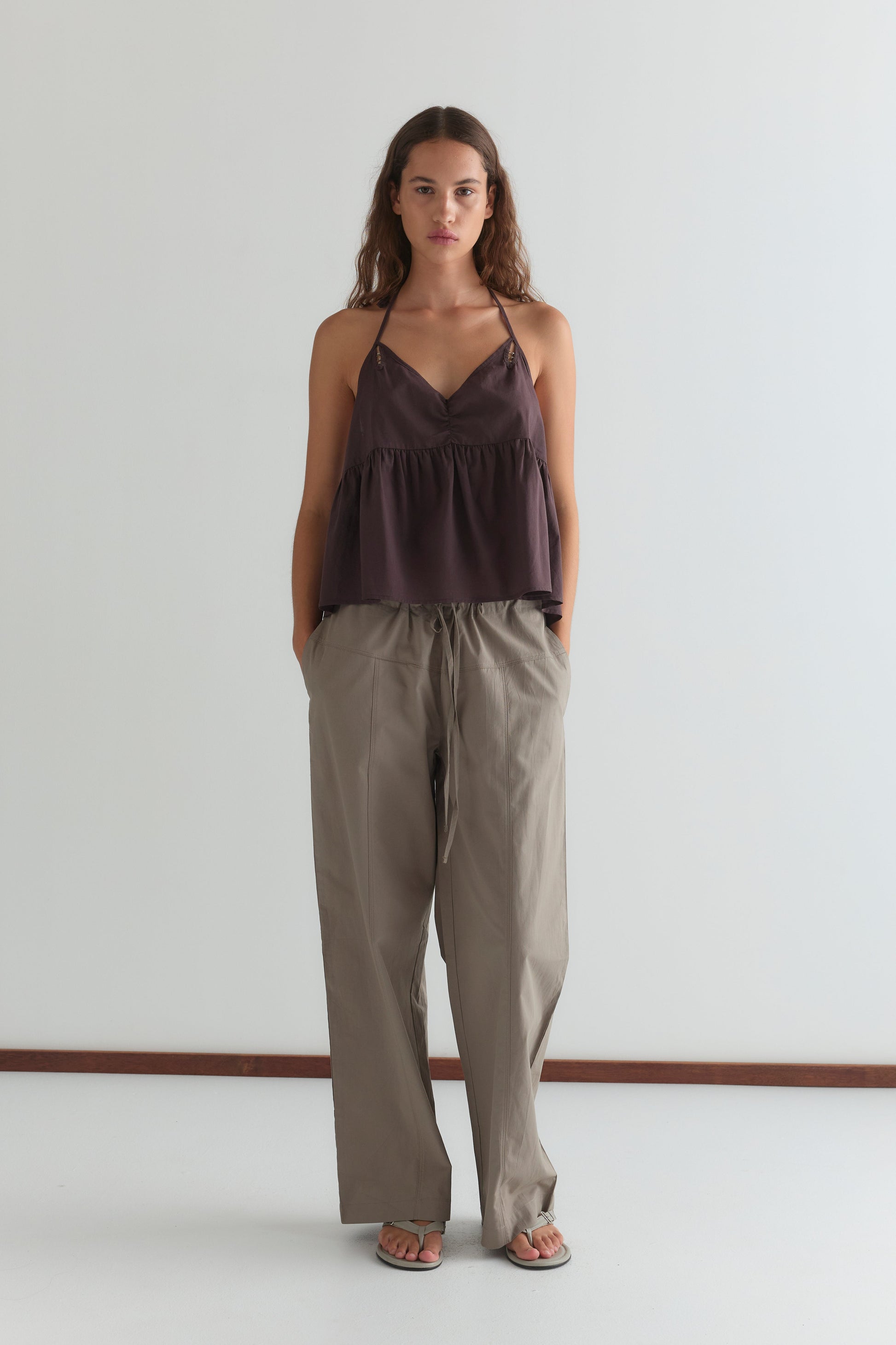 A woman with long, wavy brown hair stands against a white background, wearing the Keyhole Halter Top - Wine by Deiji Studios with adjustable straps, loose beige pants, and sandals. She has a neutral expression and hands in her pockets.