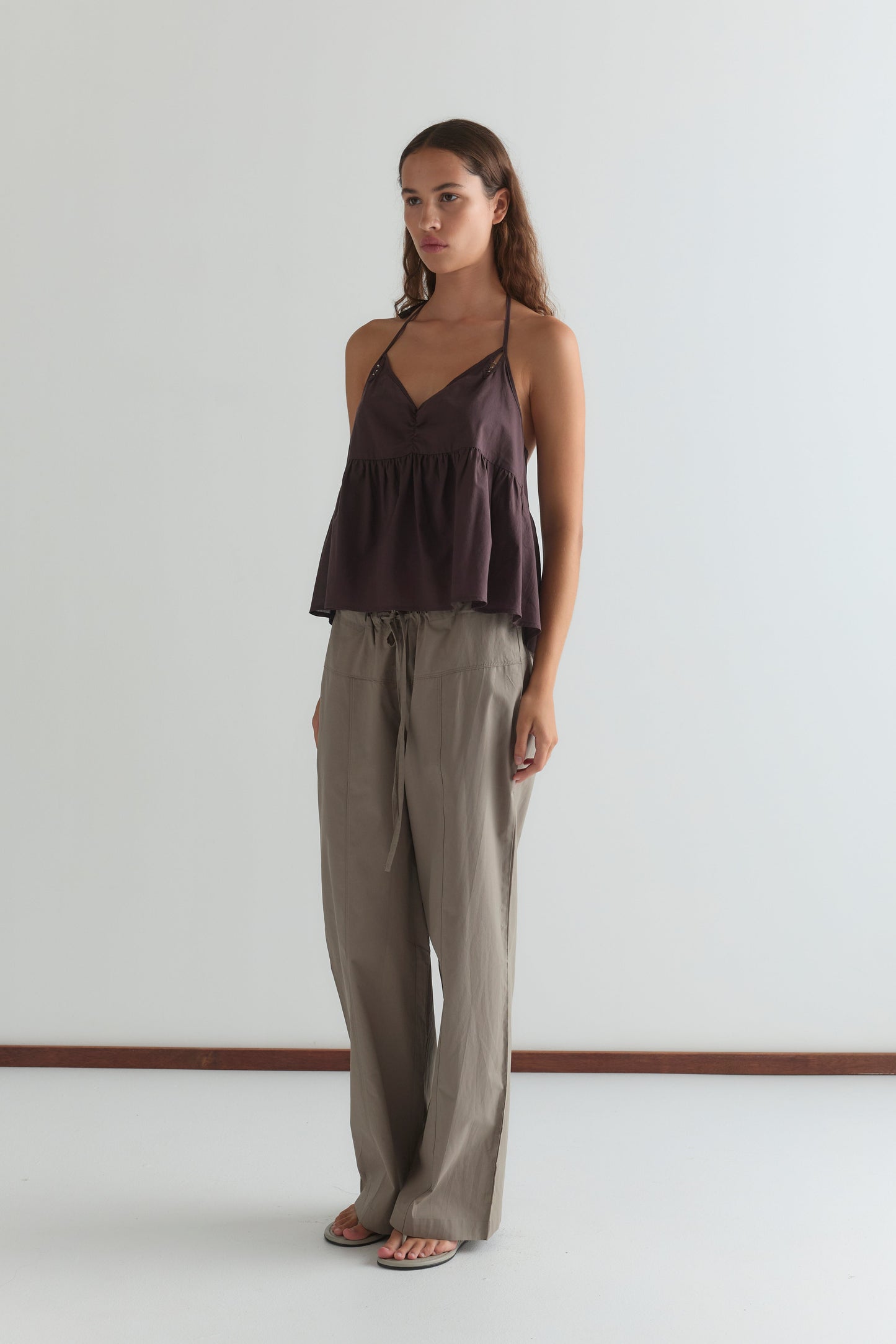 A woman with long brown hair wears the Keyhole Halter Top - Wine by Deiji Studios, featuring adjustable straps, paired with loose beige pants and flip-flops, standing against a plain white wall.