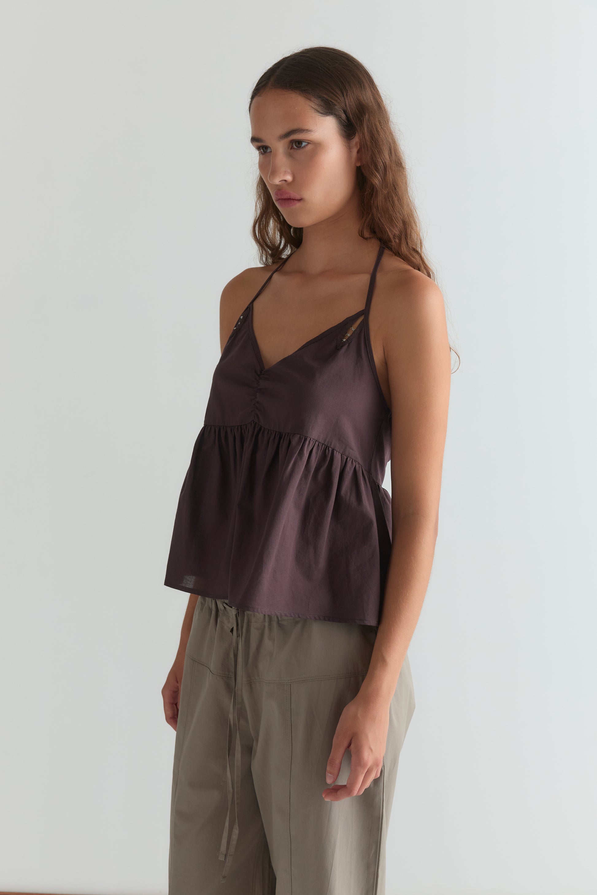 A young woman with long brown hair wears the Keyhole Halter Top - Wine by Deiji Studios, featuring adjustable straps, paired with loose beige pants, standing against a plain light background.