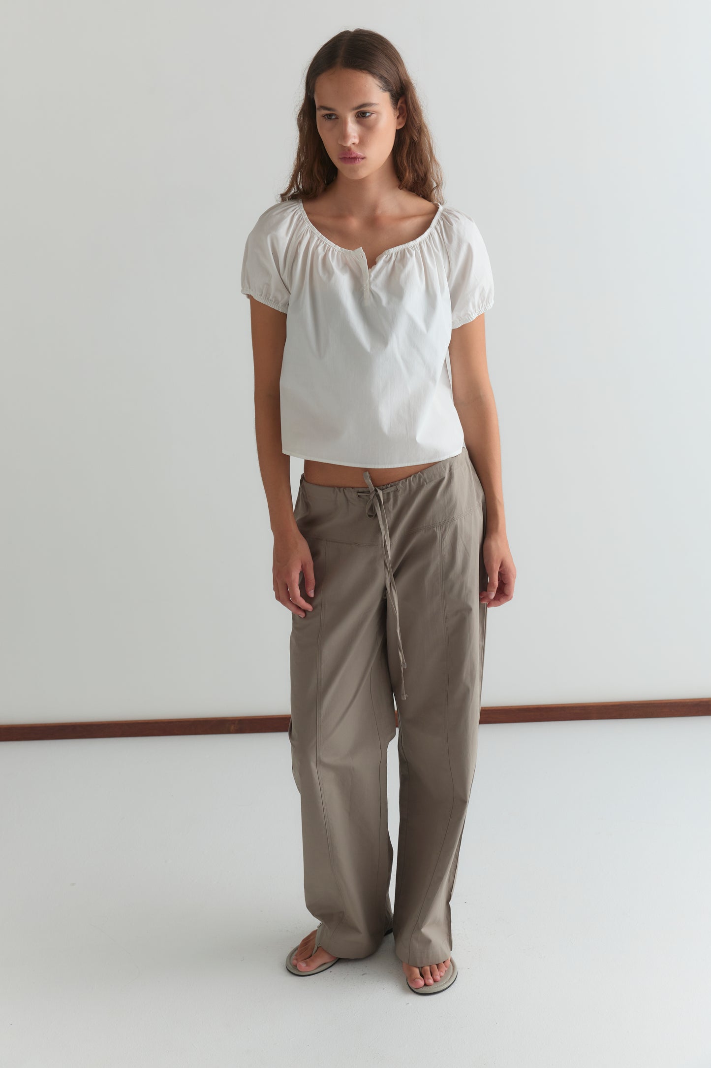 A woman with long brown hair and a neutral expression stands against a white wall, wearing the Deiji Studios Poplin T Shirt - White with puffed sleeves, loose beige drawstring pants, and sandals.
