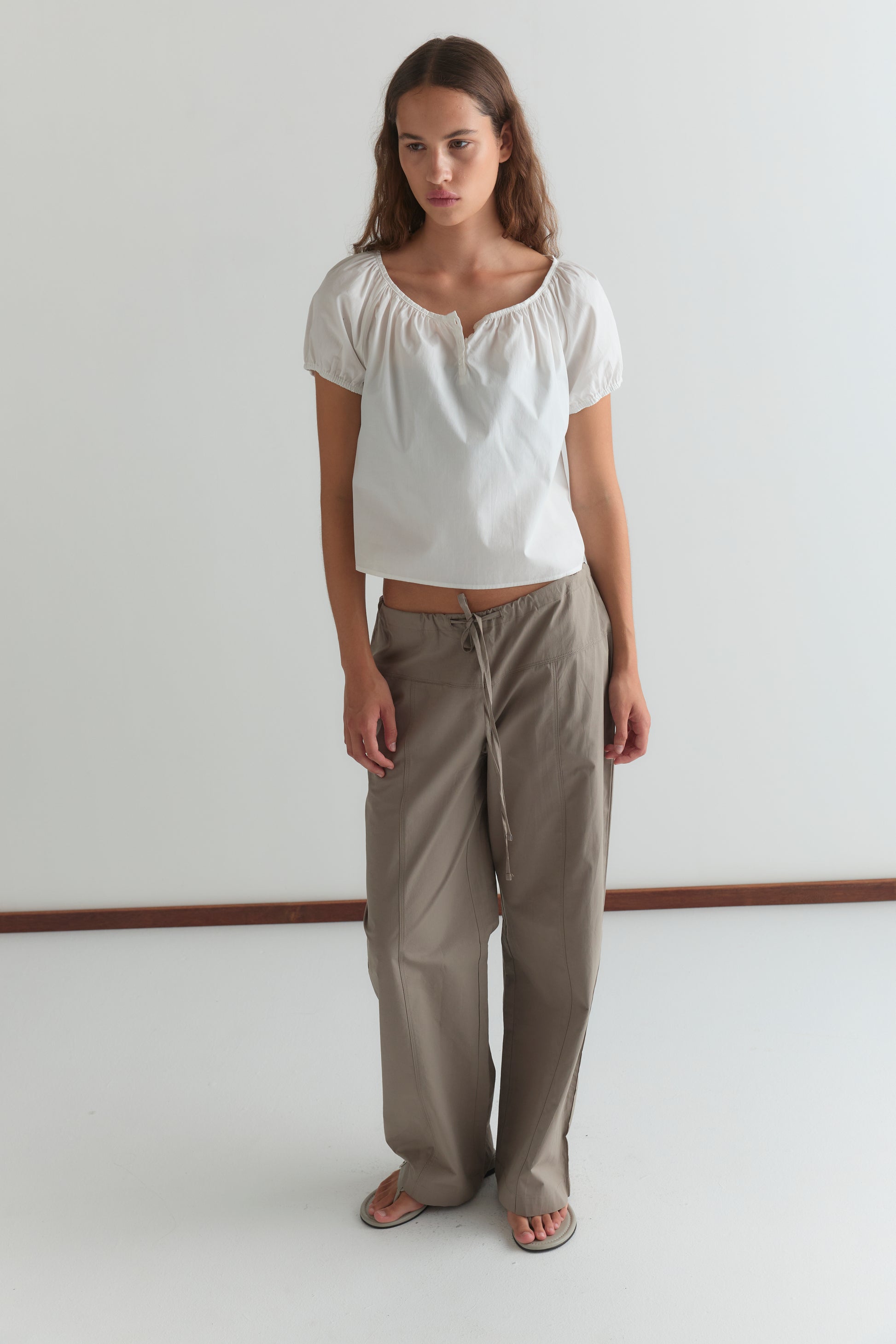 A woman with long brown hair and a neutral expression stands against a white wall, wearing the Deiji Studios Poplin T Shirt - White with puffed sleeves, loose beige drawstring pants, and sandals.