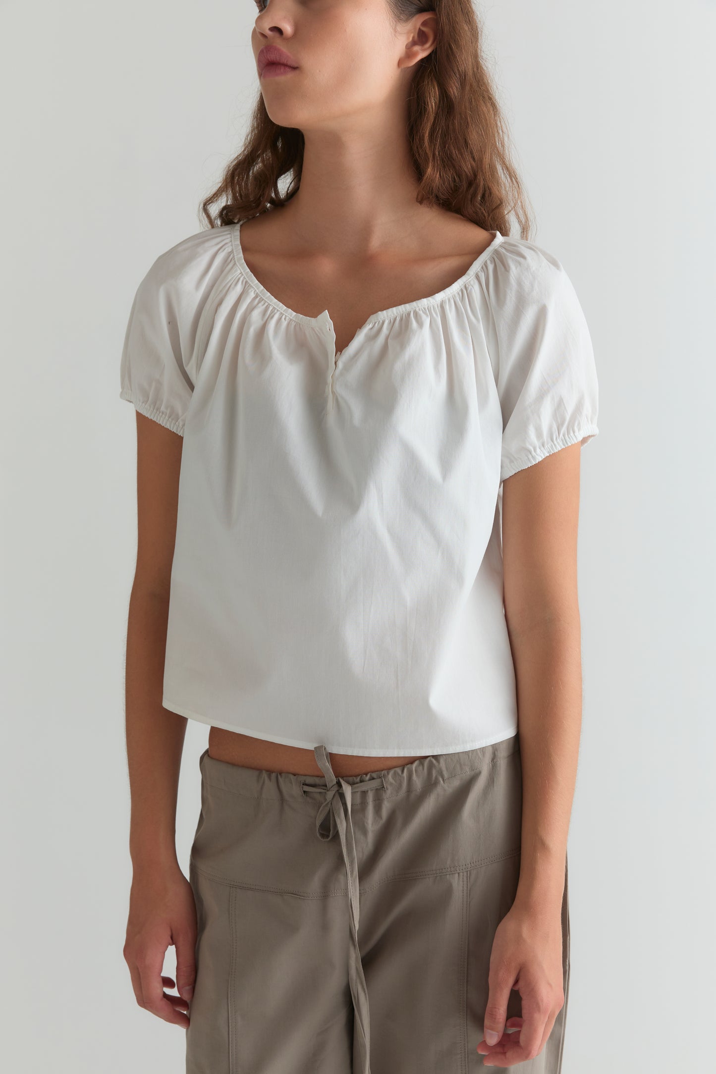 A woman stands against a plain background wearing the Deiji Studios Poplin T Shirt in white with light taupe drawstring pants. Her face is partially out of frame.