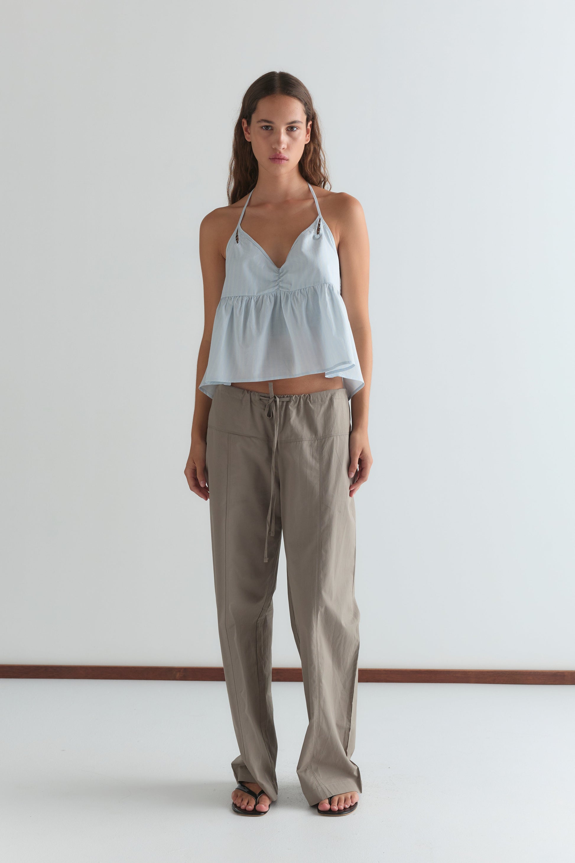 A woman with long brown hair wears the Keyhole Halter Top in Powder Blue Stripe from Deiji Studios, paired with loose beige pants and black flip-flops, standing against a plain white background.