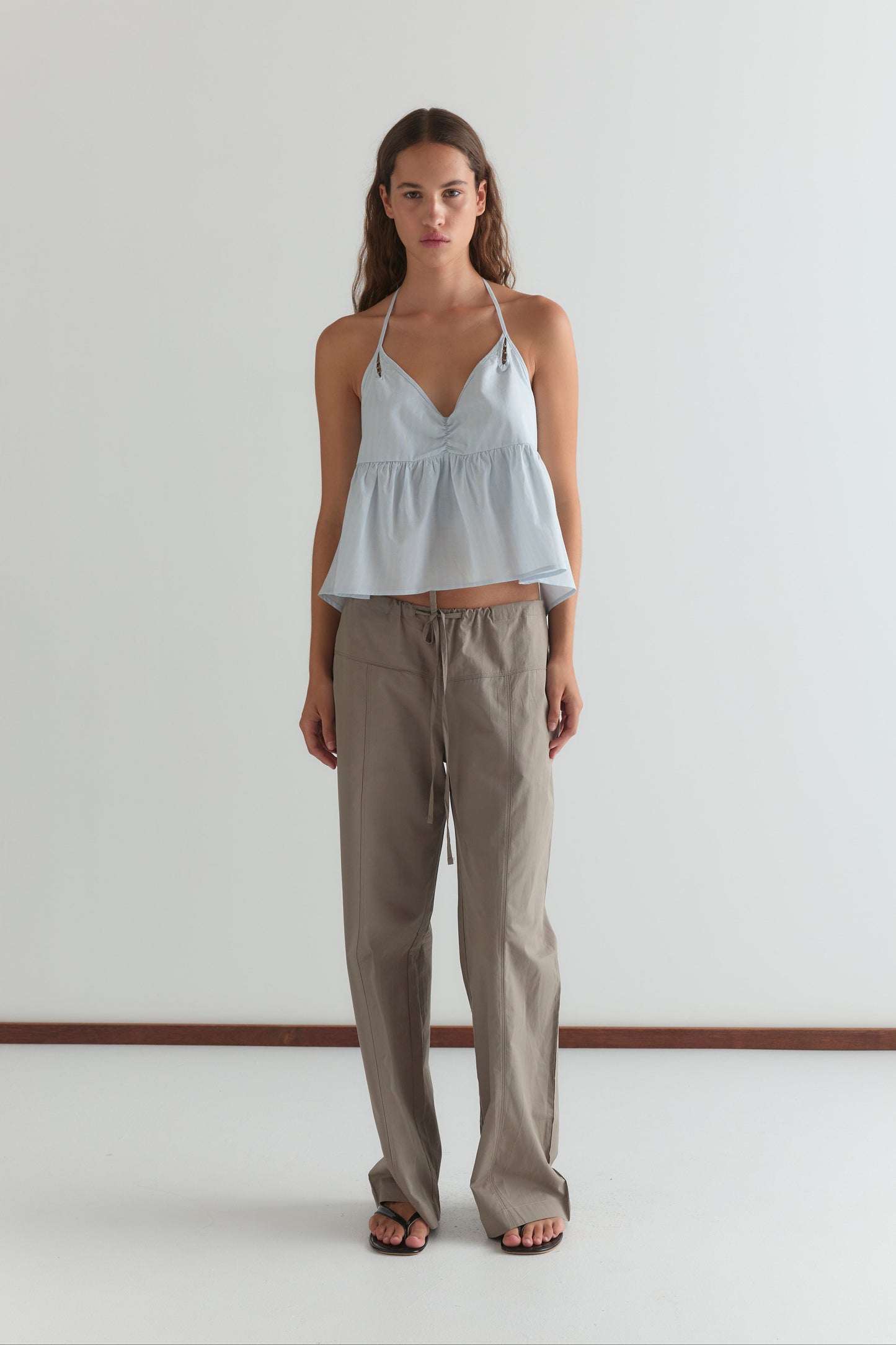 A woman with long brown hair and a neutral expression stands against a white wall, wearing a light blue v-neck sleeveless peplum top, black flip-flops, and Deiji Studios' Seam Pant in Light Khaki.