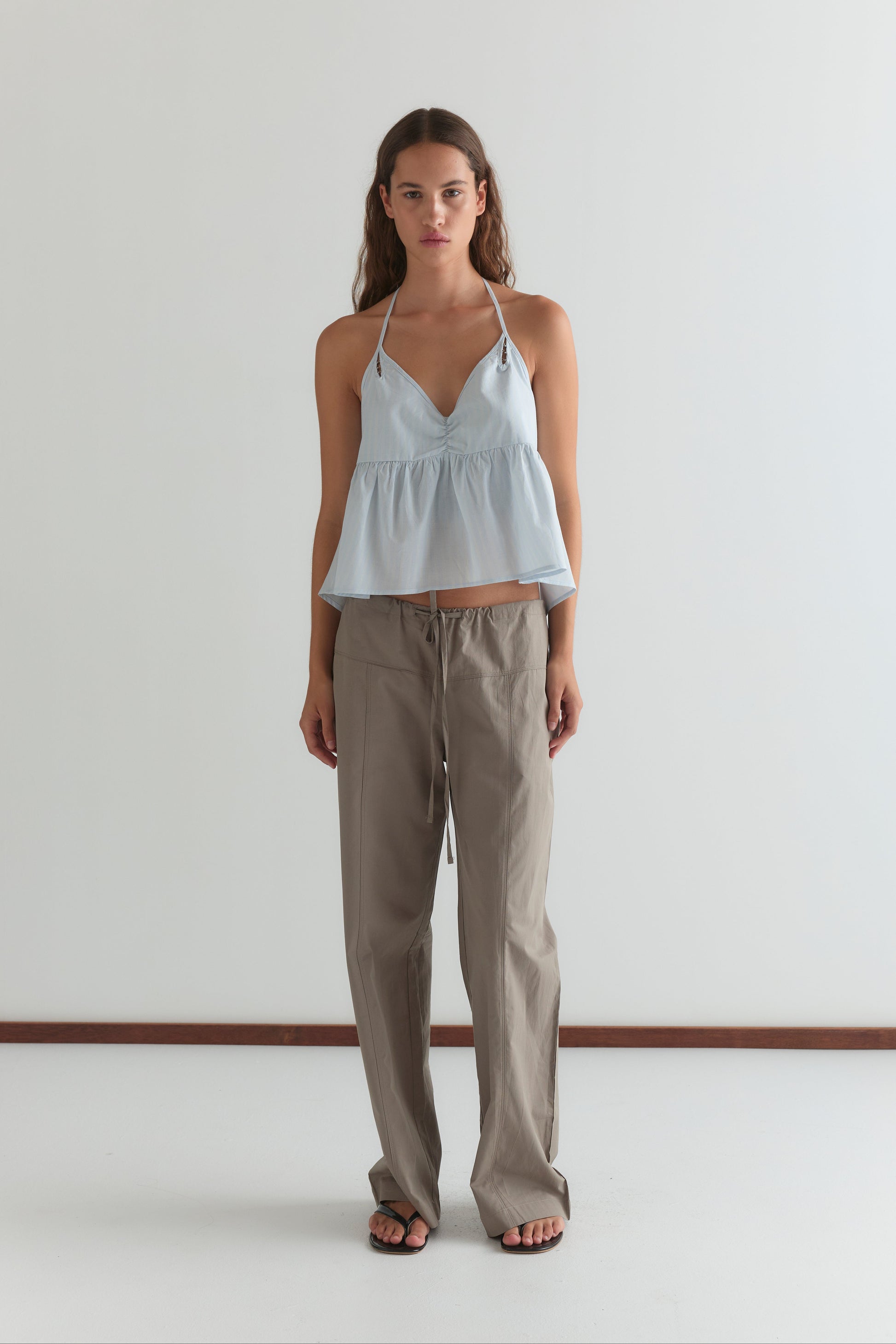 A woman with long brown hair and a neutral expression stands against a white wall, wearing a light blue v-neck sleeveless peplum top, black flip-flops, and Deiji Studios' Seam Pant in Light Khaki.