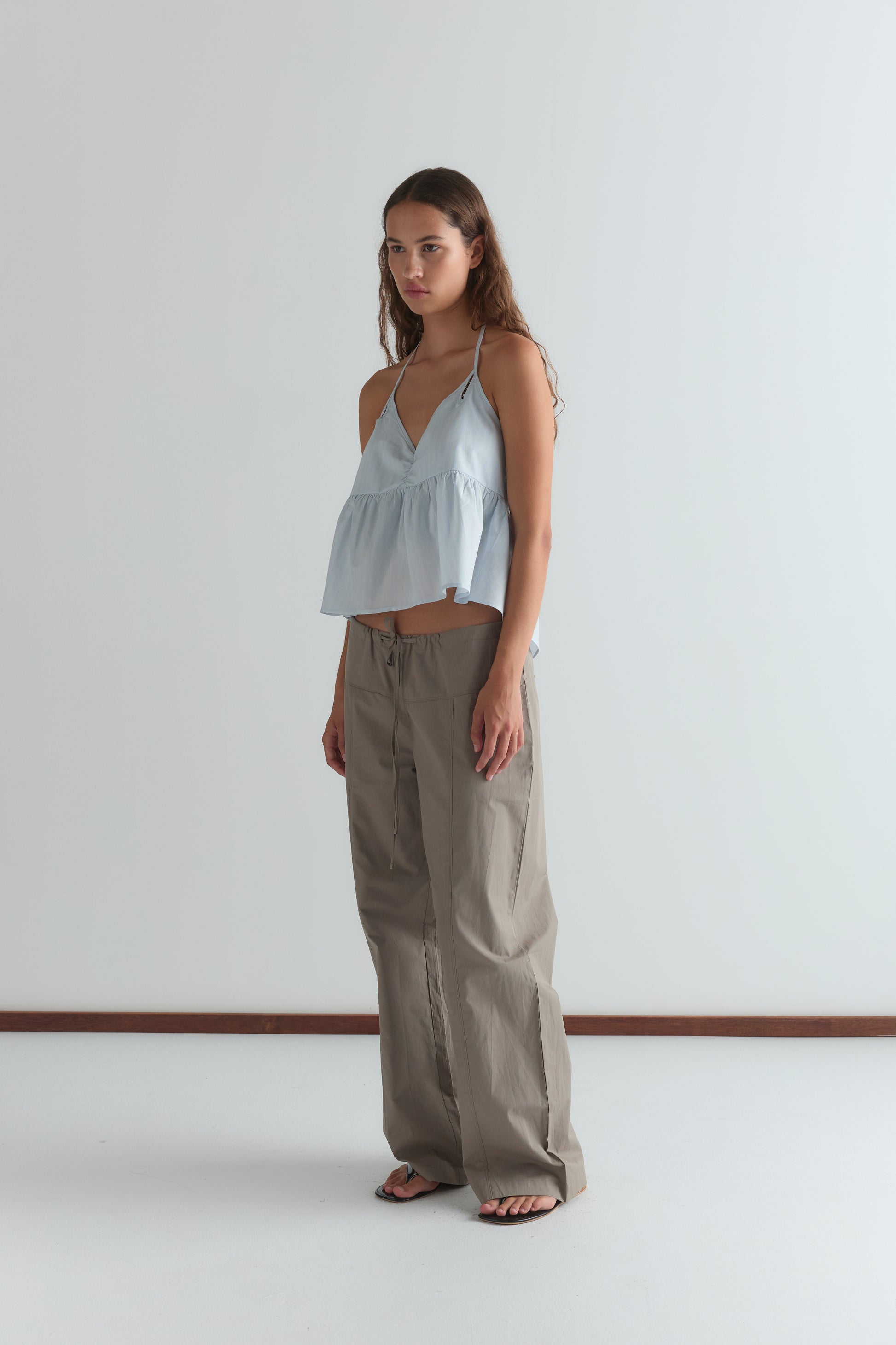 A woman with long brown hair stands barefoot indoors against a white wall, wearing the Keyhole Halter Top - Powder Blue Stripe and loose, wide-legged khaki pants.