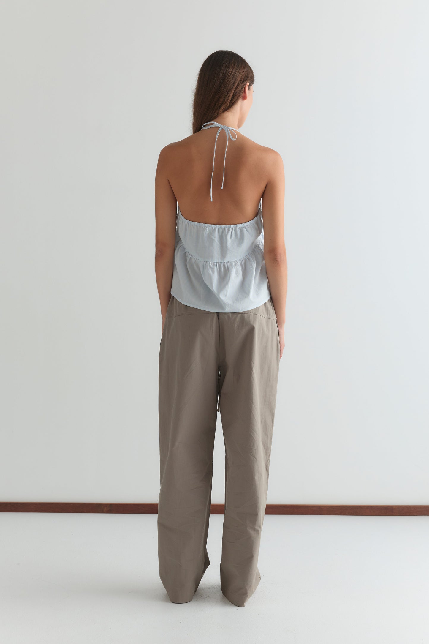 A woman stands facing away, wearing a light blue halter-top and loose, high-waisted Seam Pant - Light Khaki from Deiji Studios, against a plain white wall.
