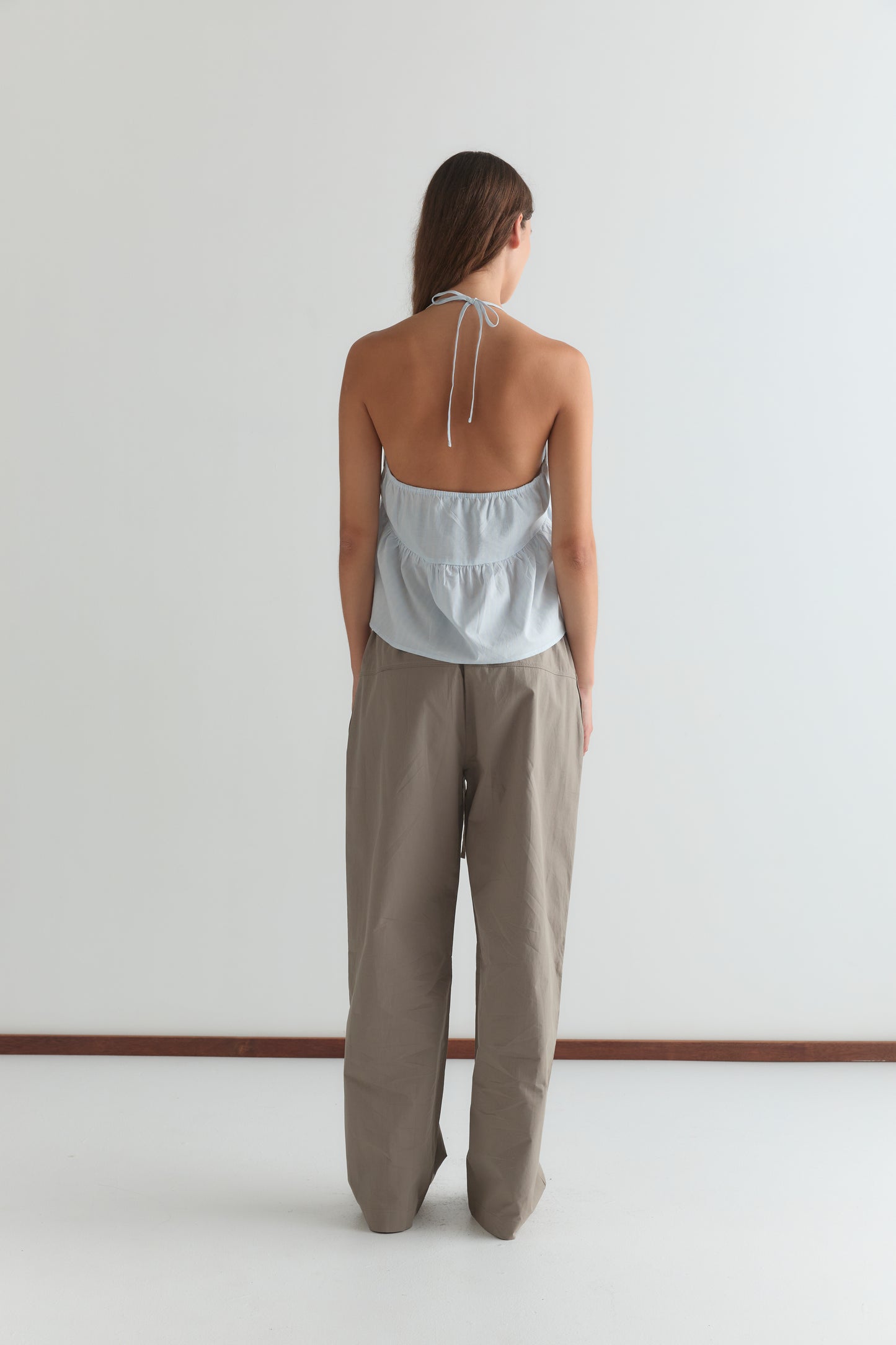 A person with long brown hair faces away, wearing the Keyhole Halter Top - Powder Blue Stripe and loose taupe pants against a plain white background.