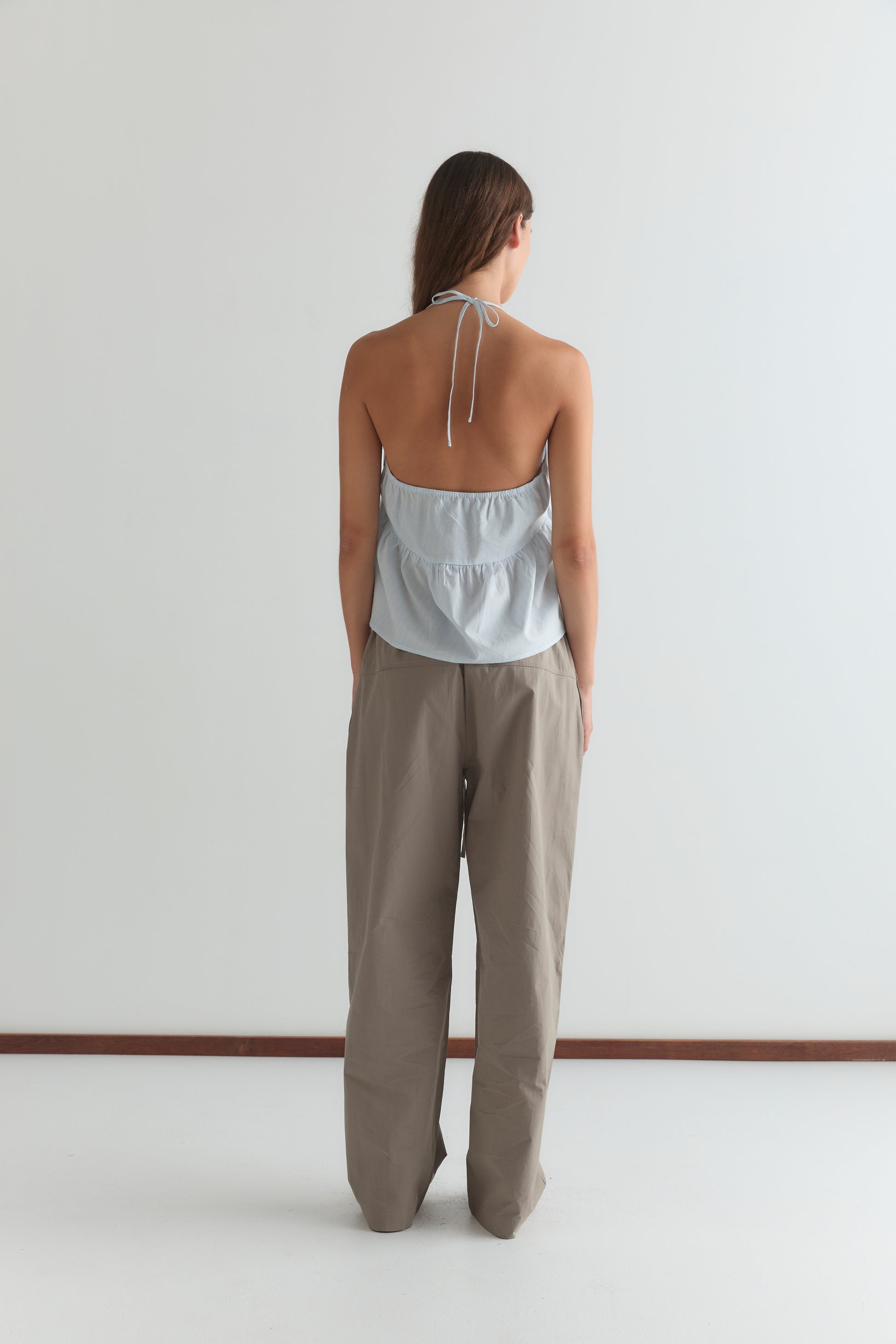 A person with long brown hair faces away, wearing the Keyhole Halter Top - Powder Blue Stripe and loose taupe pants against a plain white background.