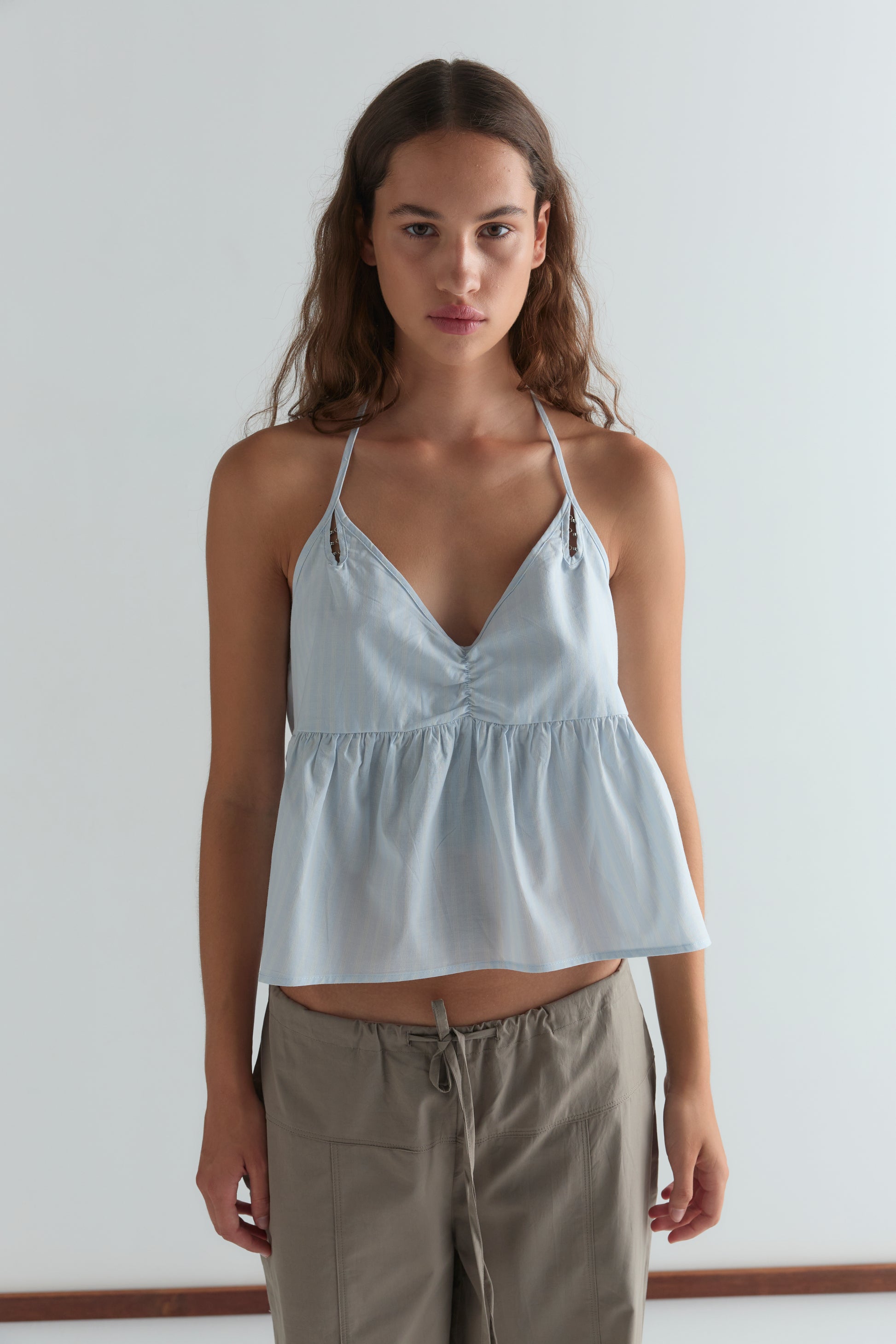 A woman with long brown hair wears a light blue, sleeveless V-neck top with thin straps and the Seam Pant in Light Khaki from Deiji Studios, standing against a plain white background.