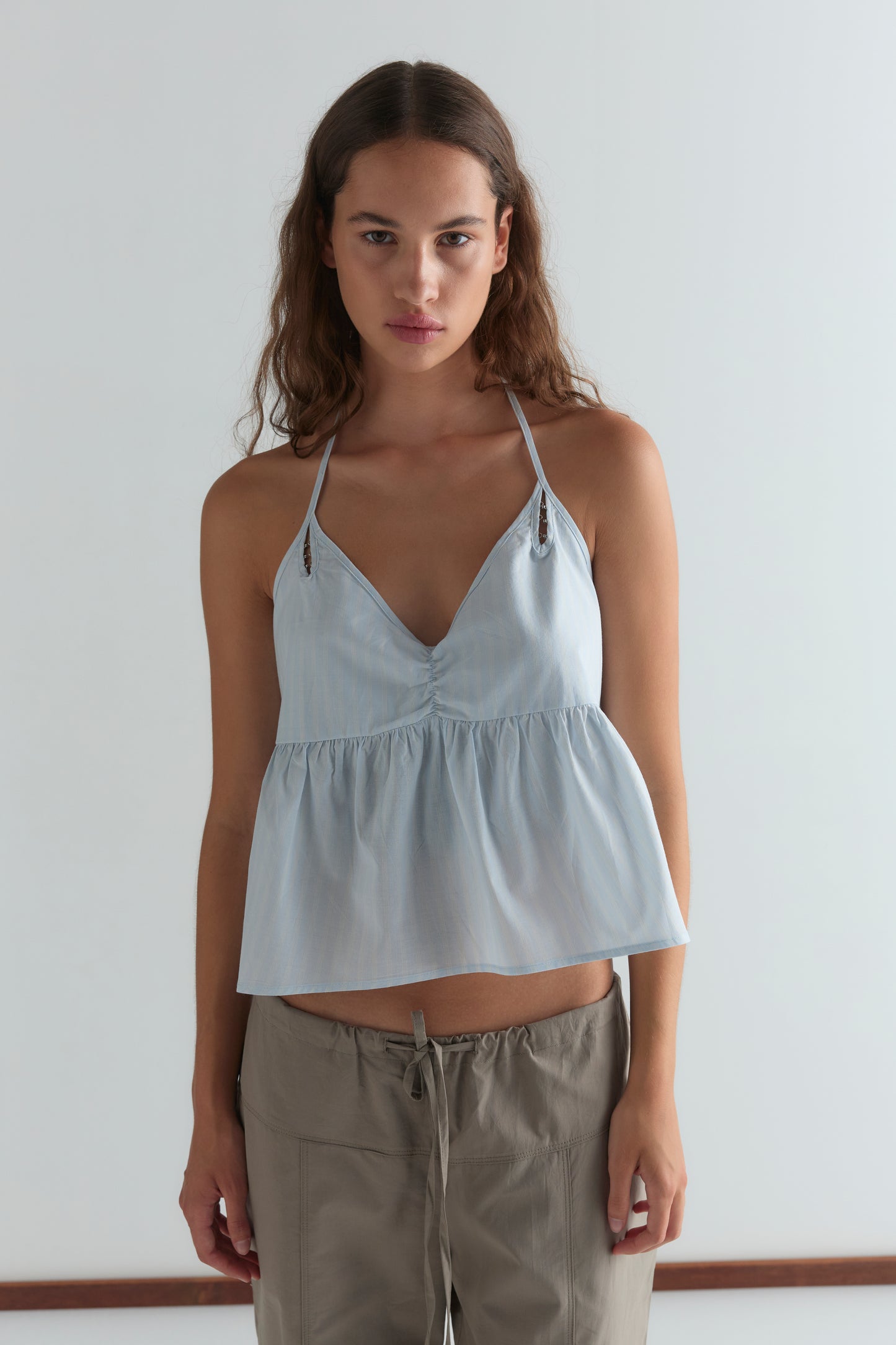 A woman with long brown hair wears the Deiji Studios Keyhole Halter Top in Powder Blue Stripe and khaki drawstring pants, standing against a plain white background.