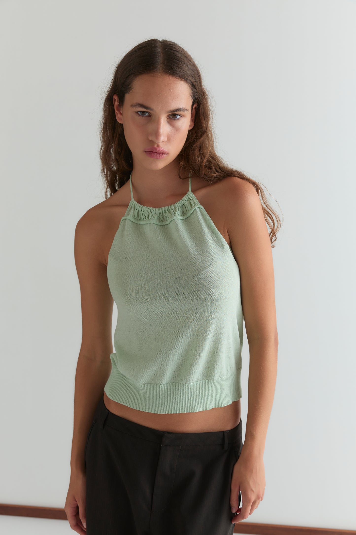 A young woman with long brown hair wears the Deiji Studios Drawcord Knit Top in Mint, featuring an adjustable ruched neckline, paired with dark pants. She stands against a white background, looking at the camera with a neutral expression.