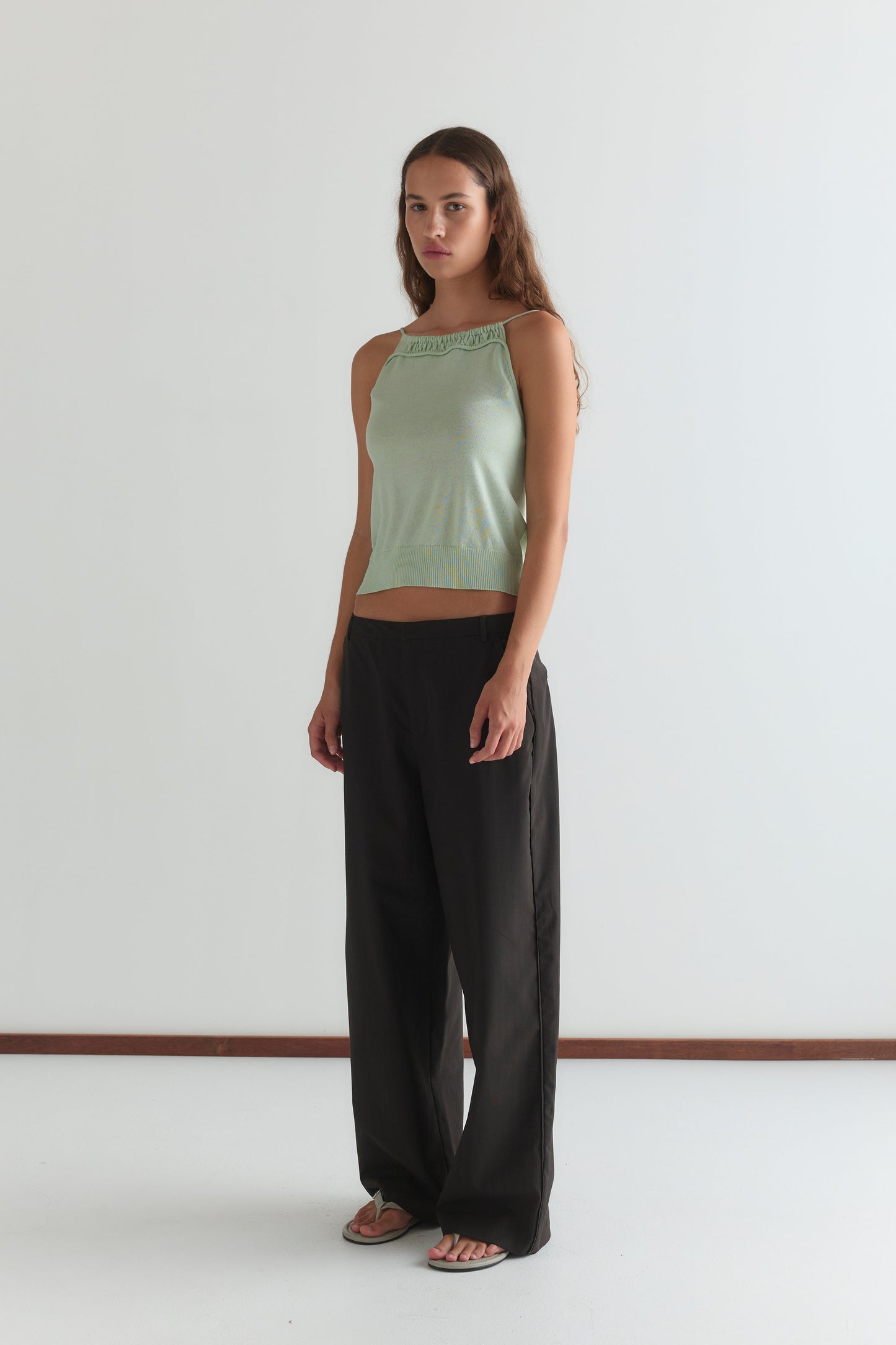 A woman with long brown hair stands indoors against a plain white wall, wearing the Drawcord Knit Top - Mint by Deiji Studios, loose black pants, and white sandals. She has a neutral expression.