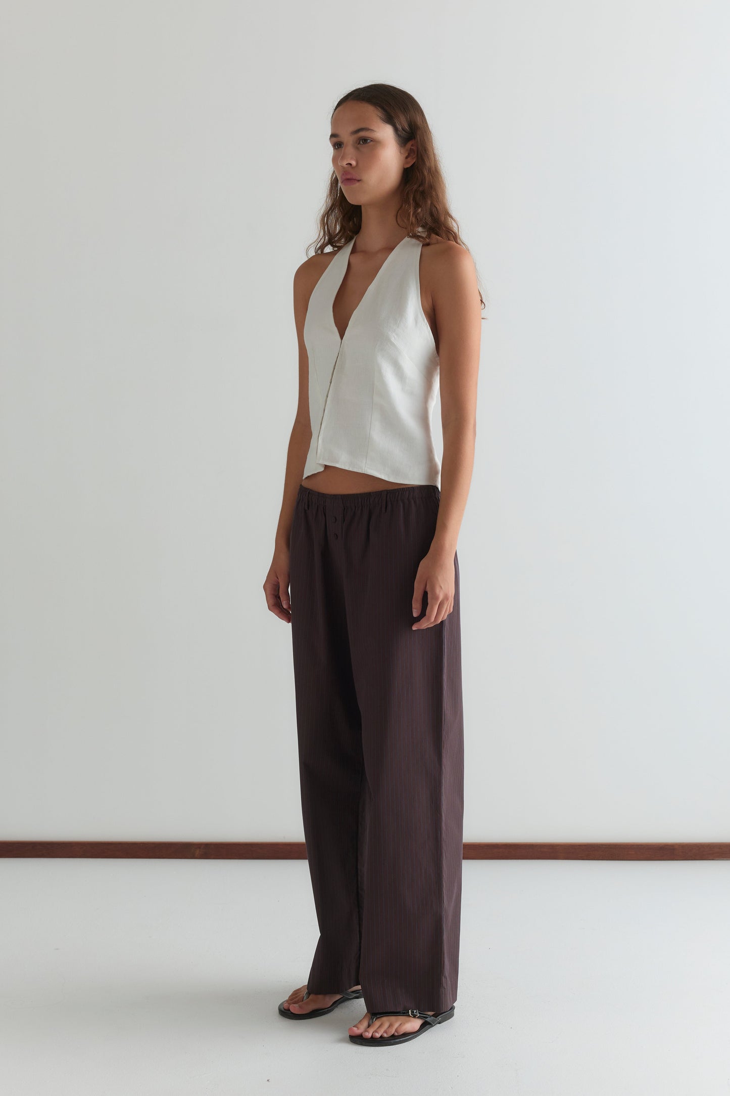 A woman with long wavy hair stands against a white wall, wearing the Double Button Pant - Brown Stripe and a sleeveless white vest, paired with black sandals. She has a neutral expression and her arms rest relaxed at her sides.