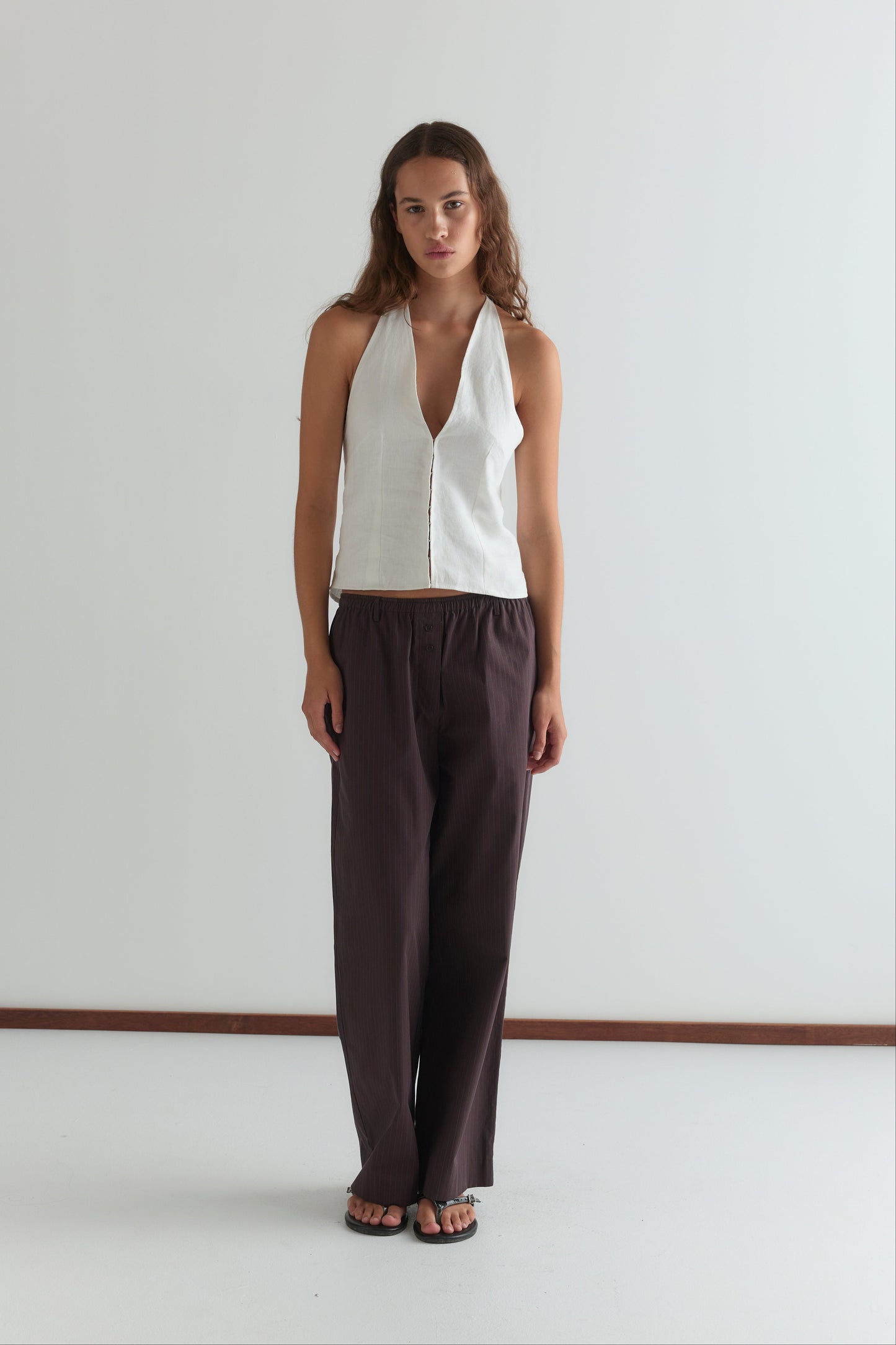 A woman with long hair stands against a white wall, wearing Deiji Studios’ Close Halter Top in white—a sleeveless halter style with a deep V neckline—paired with loose dark brown wide-leg pants. She gazes ahead with a neutral expression.