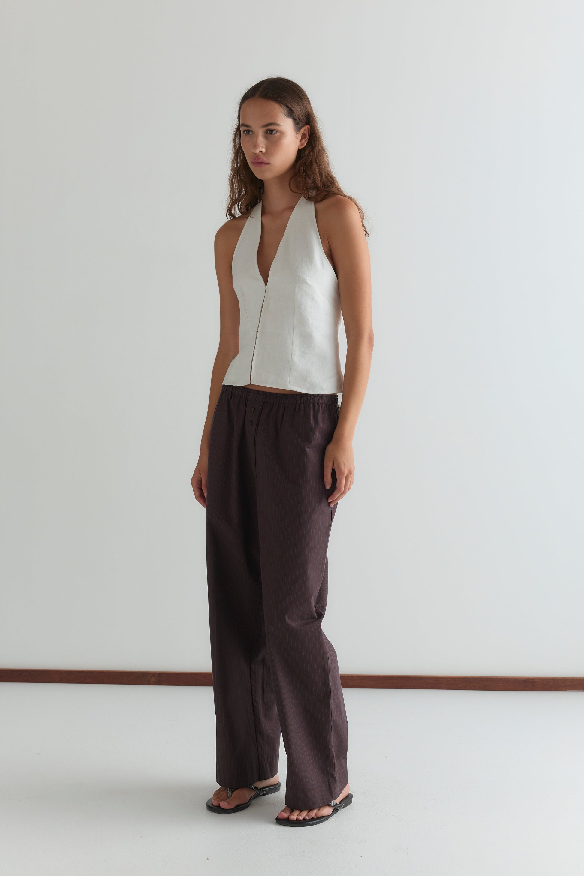A woman with long, wavy hair and a neutral expression stands against a plain white wall wearing a sleeveless white V-neck top, black sandals, and Deiji Studios’ Double Button Pant - Brown Stripe.
