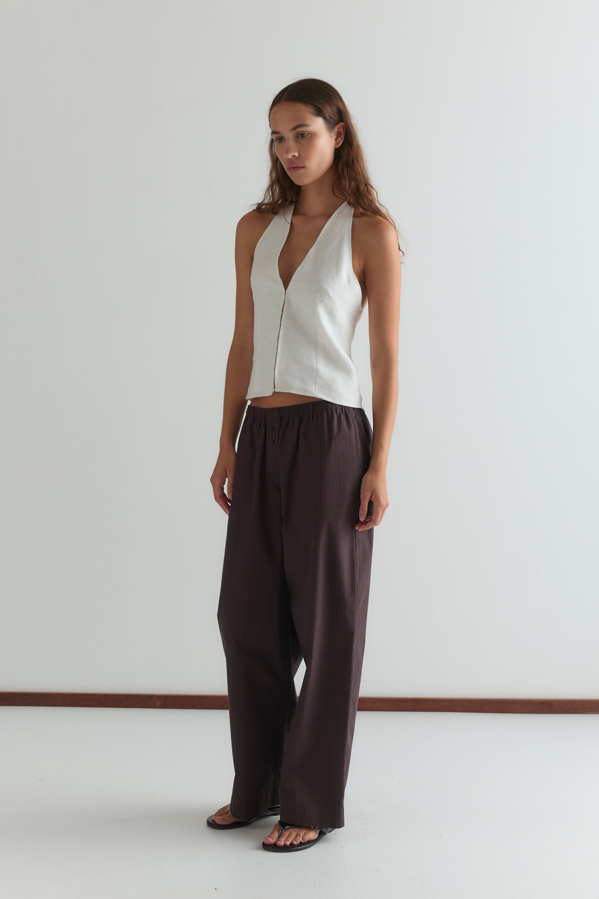 A woman with long brown hair and a neutral expression stands in a plain room wearing a sleeveless white V-neck top, black sandals, and Deiji Studios’ Double Button Pant - Brown Stripe.