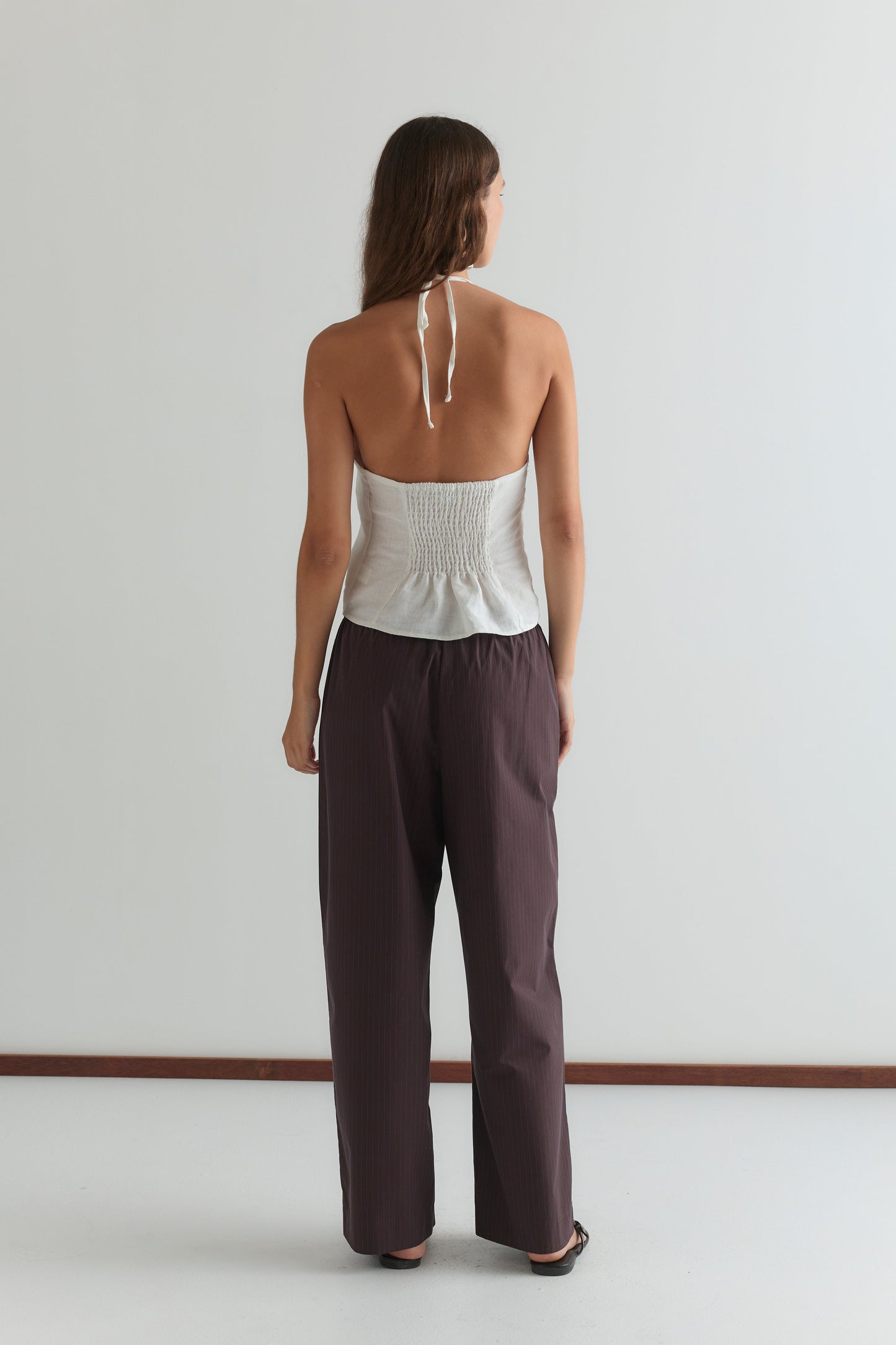 A woman with long brown hair faces away, barefoot on a white floor against a plain light wall, wearing the Close Halter Top - White by Deiji Studios with a smocked back and loose dark brown pants.