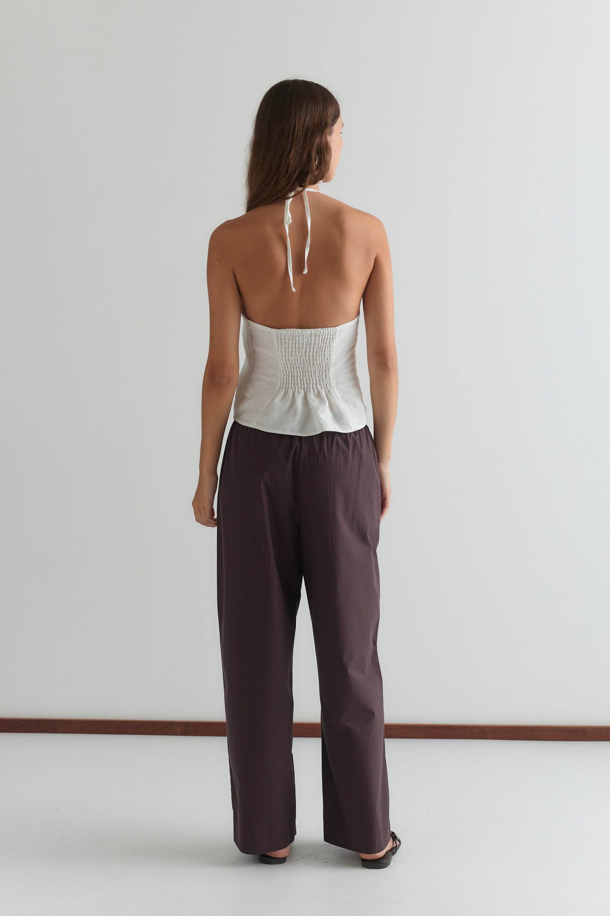 A woman with long brown hair faces away, barefoot on a white floor against a plain light wall, wearing the Close Halter Top - White by Deiji Studios with a smocked back and loose dark brown pants.