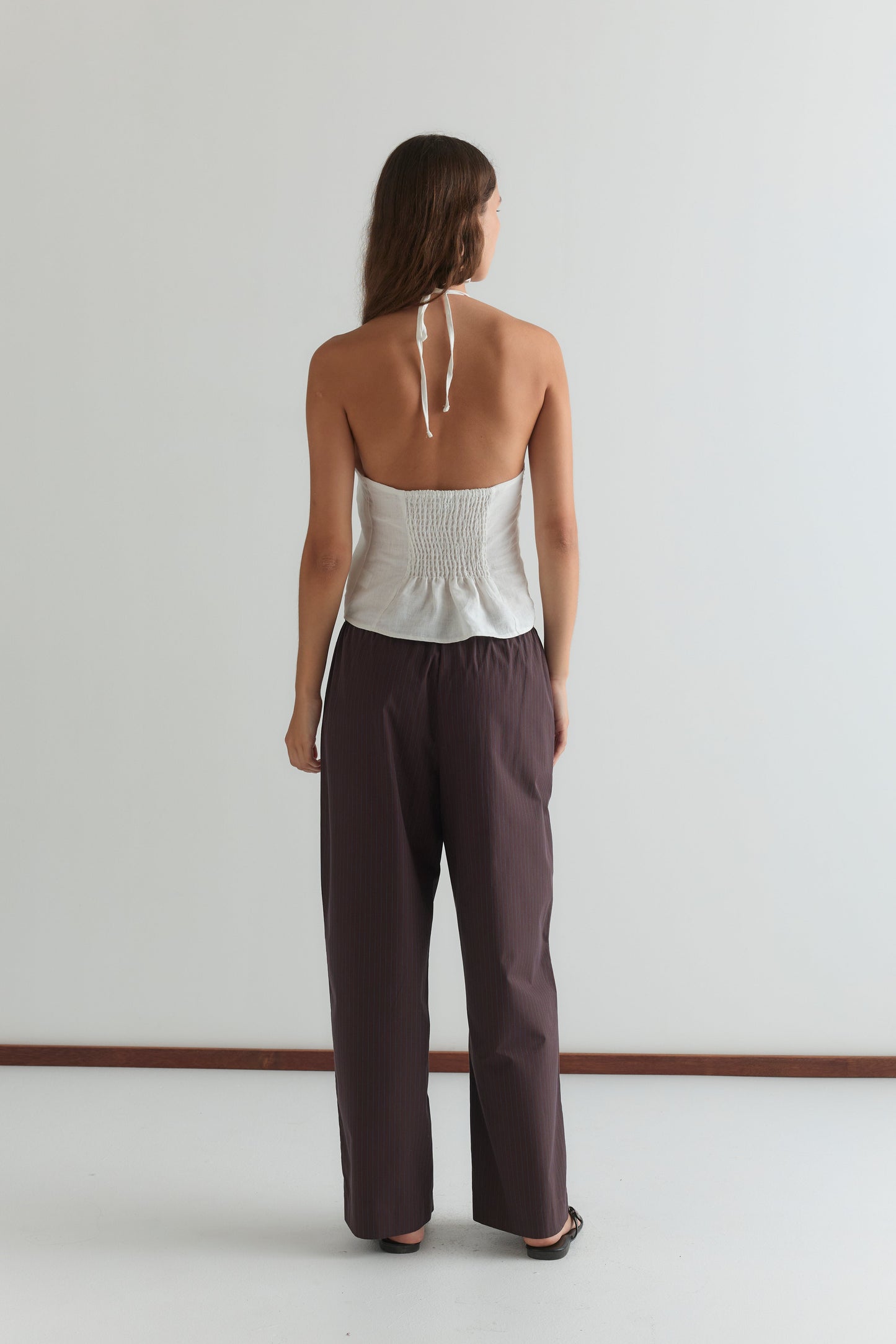 A woman stands facing away from the camera, wearing a white strapless Deiji Studios top and the Double Button Pant - Brown Stripe in a minimal, light-colored room.
