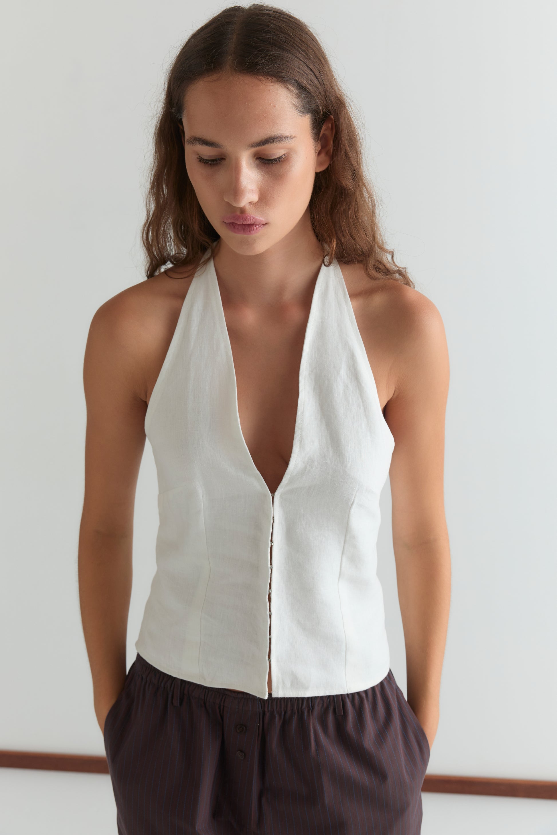 A woman with long brown hair wears the Close Halter Top, a sleeveless white halter style featuring a deep neckline, paired with dark loose-fitting pants. She stands indoors against a plain light background, looking down neutrally.