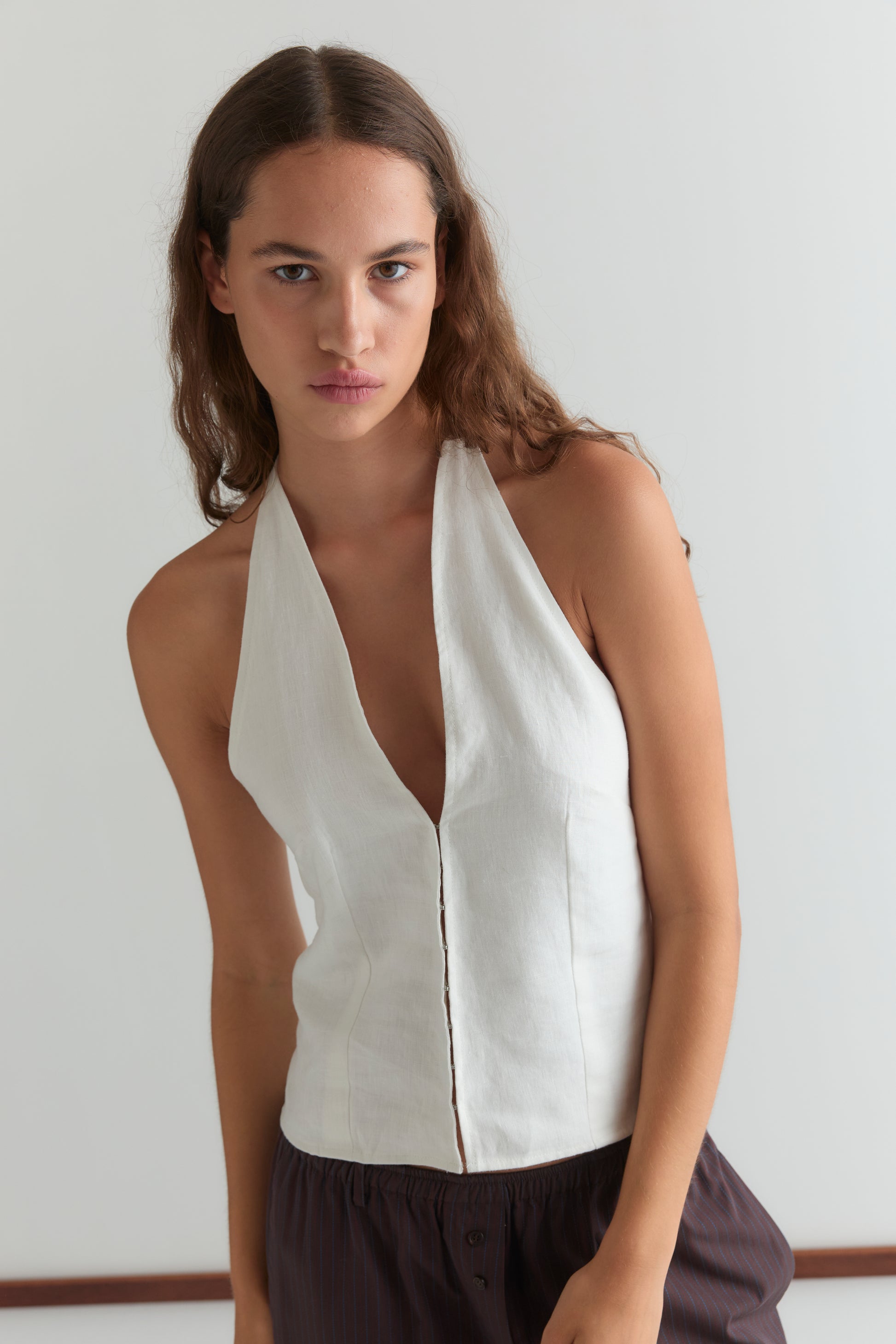 A woman with long brown hair wears the Close Halter Top - White, a sleeveless deep V-neck style, paired with dark pants. She stands against a plain light background, looking directly at the camera with a neutral expression.