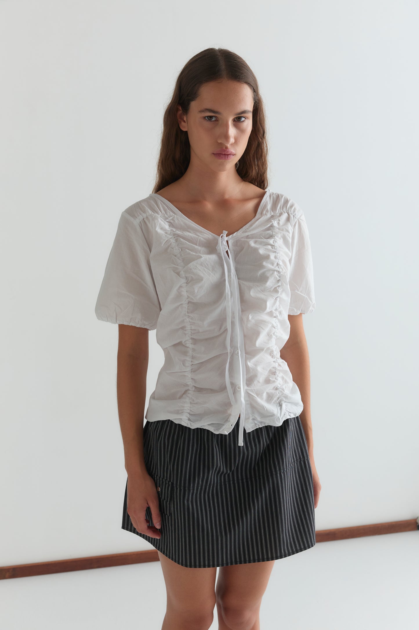 A woman with long brown hair wears the Crinkle Top - White and a black pinstriped skirt, standing against a plain white background and looking at the camera.