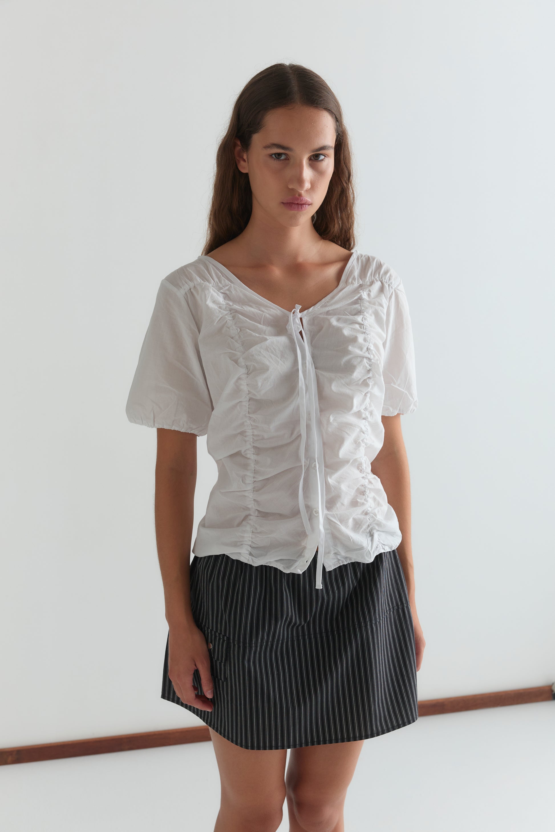 A woman with long brown hair wears the Crinkle Top - White and a black pinstriped skirt, standing against a plain white background and looking at the camera.