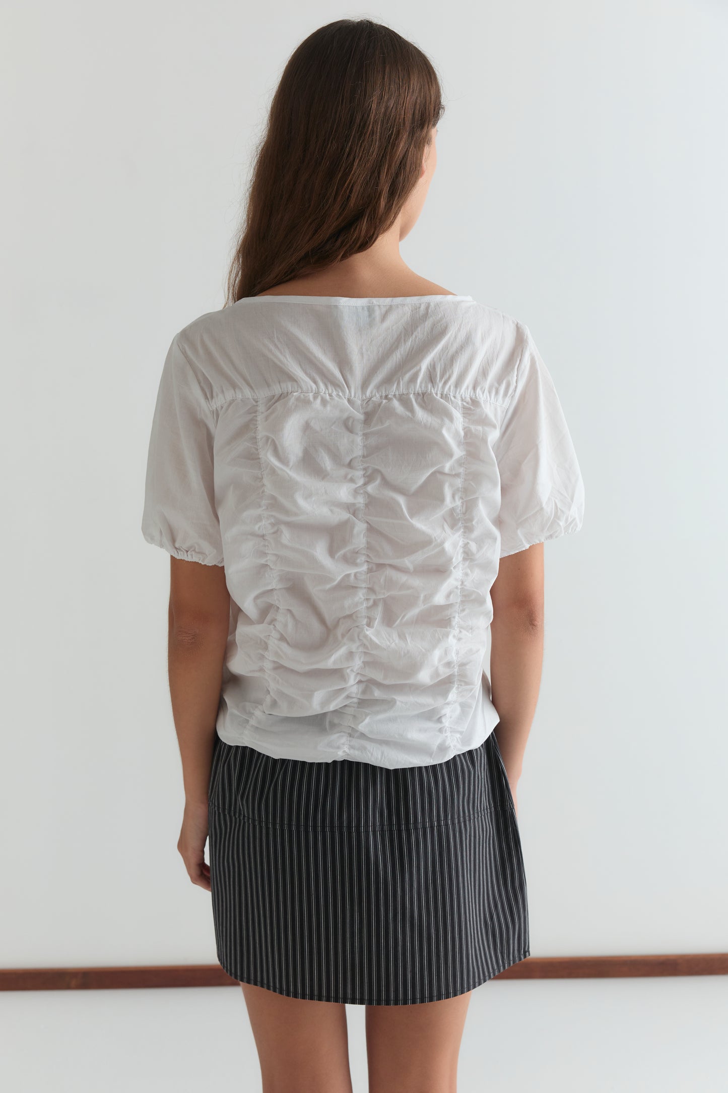 A woman with long brown hair, seen from behind, wears the Crinkle Top - White by Deiji Studios and a black pinstripe skirt, standing against a plain white background.