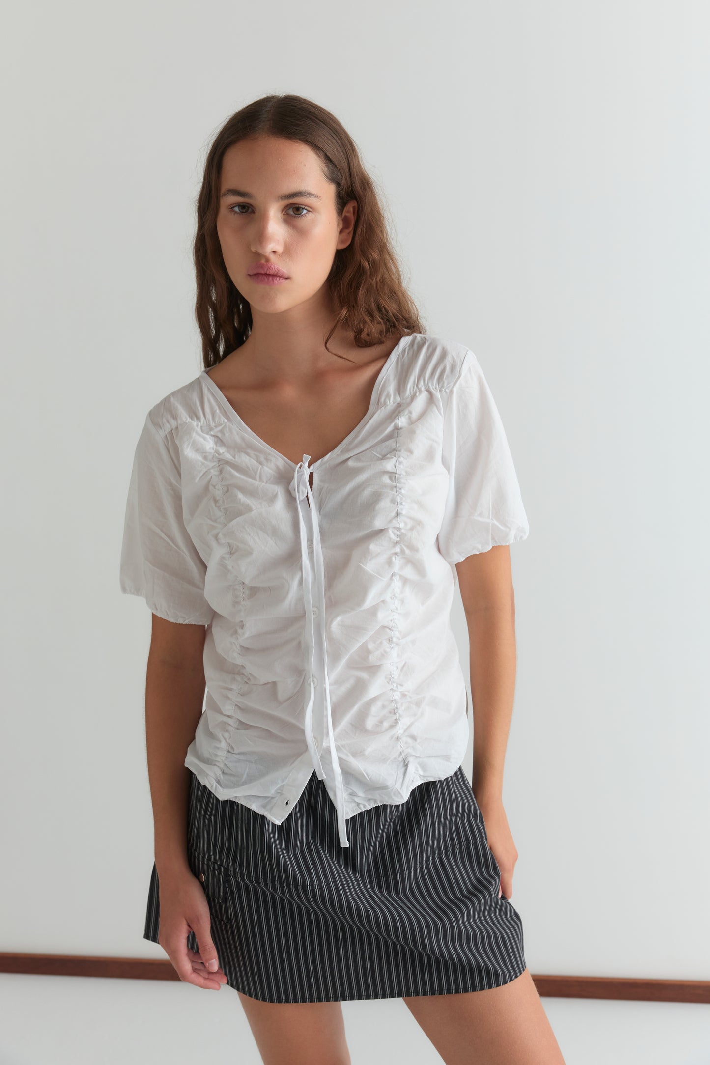 A woman with long brown hair wears the Deiji Studios Crinkle Top - White and a black pinstriped mini skirt, standing against a plain white background.