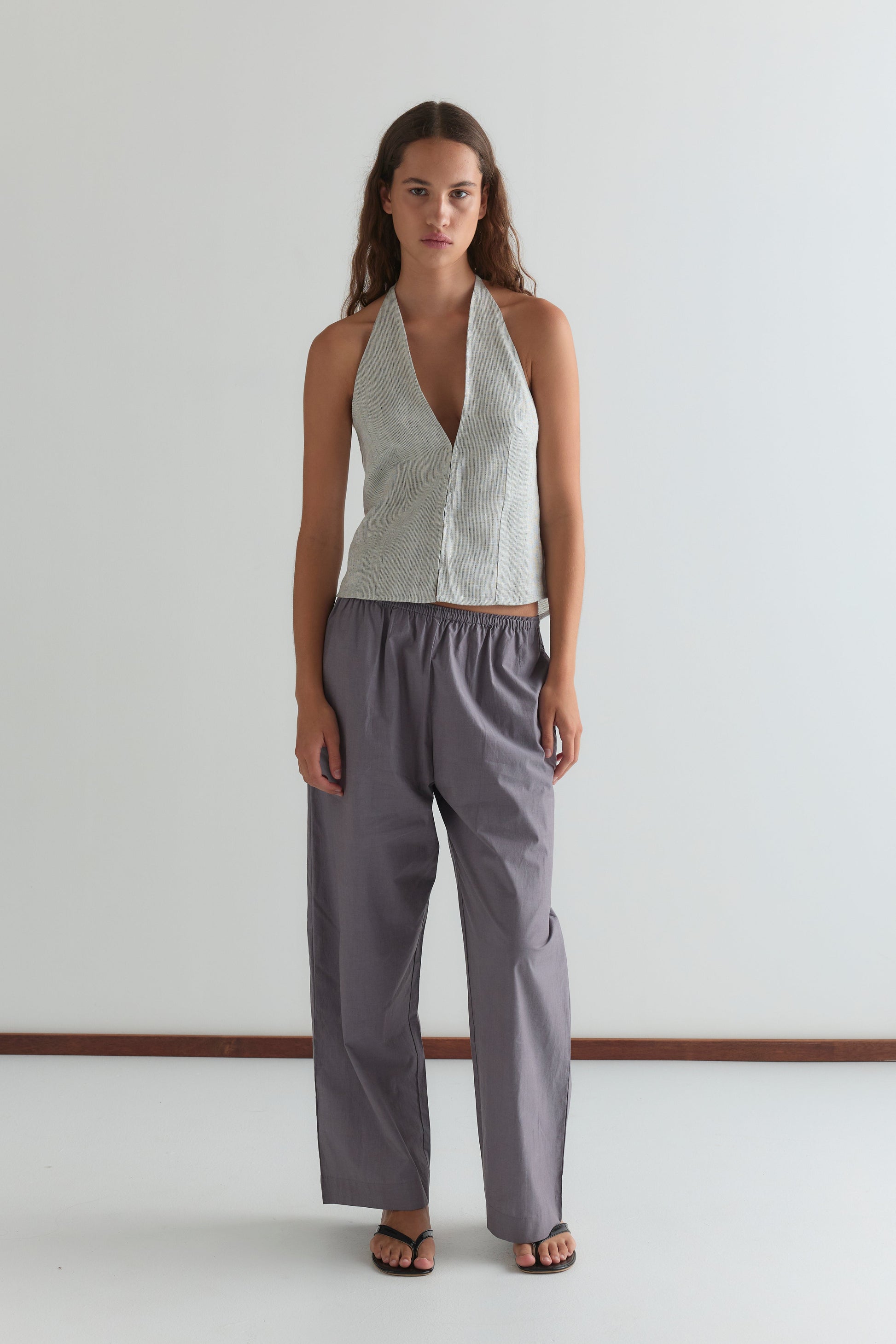 A woman with long brown hair and black sandals stands against a plain white wall, wearing Deiji Studios' Close Halter Top - Fine Stripe with a deep V neckline and loose, high-waisted gray pants.