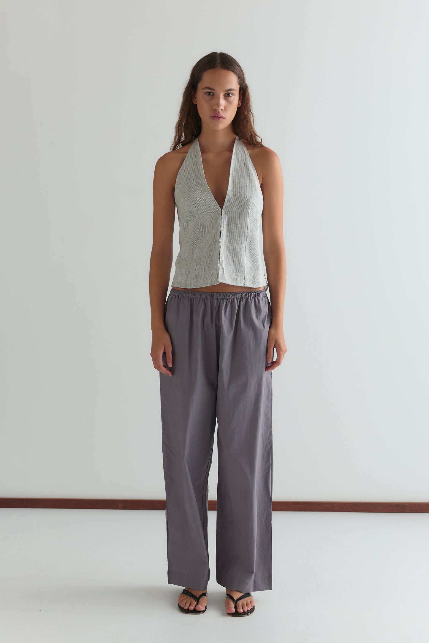 A woman with long brown hair and a neutral expression stands against a white wall, wearing the Ease Trouser—loose, high-waisted gray pants—paired with a sleeveless light gray halter top and black sandals.