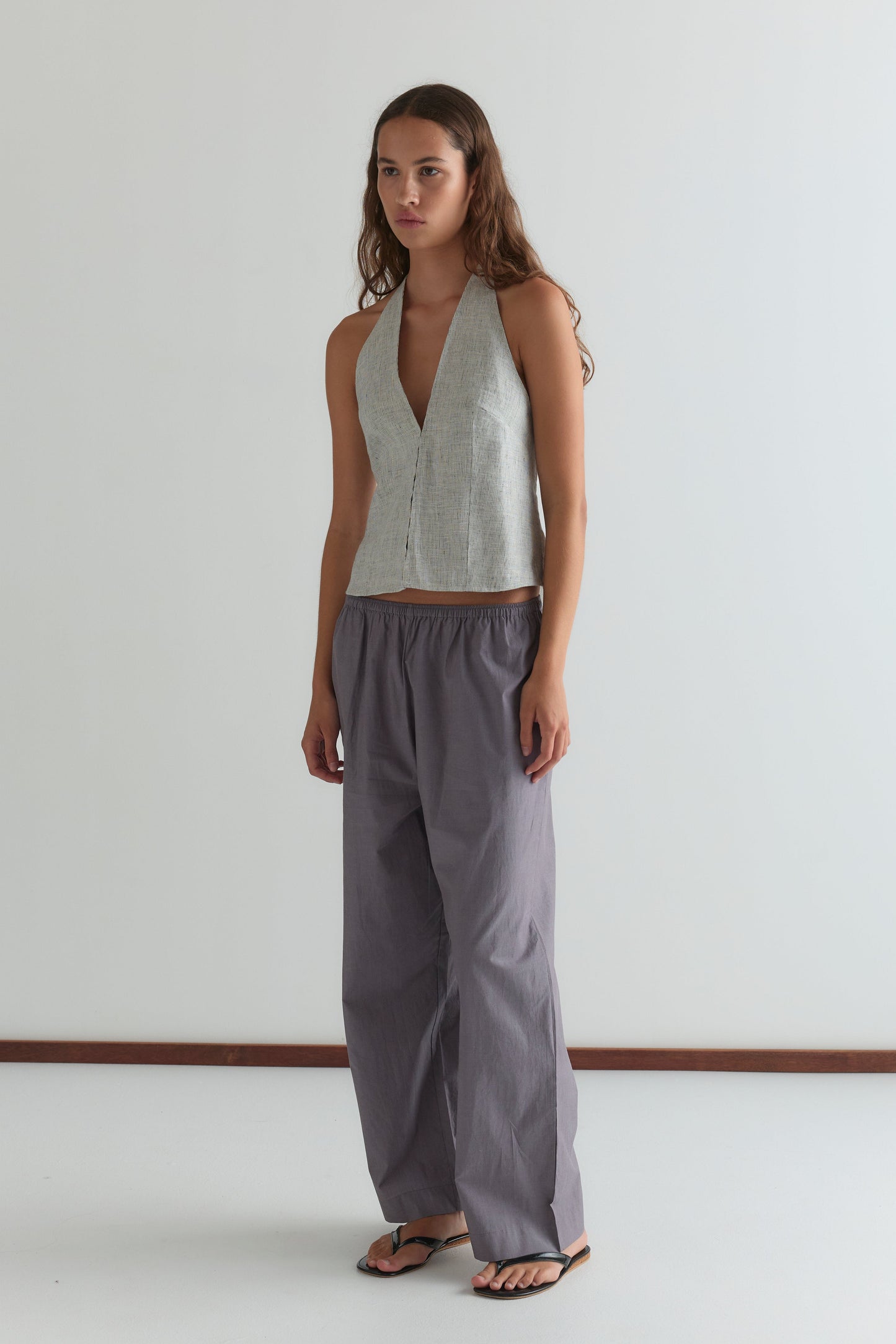 A woman stands against a white wall in a silver halter top, black flip-flops, and Deiji Studios Ease Trouser - Currant. She has long brown hair, an ankle-length pant silhouette, and a neutral expression.