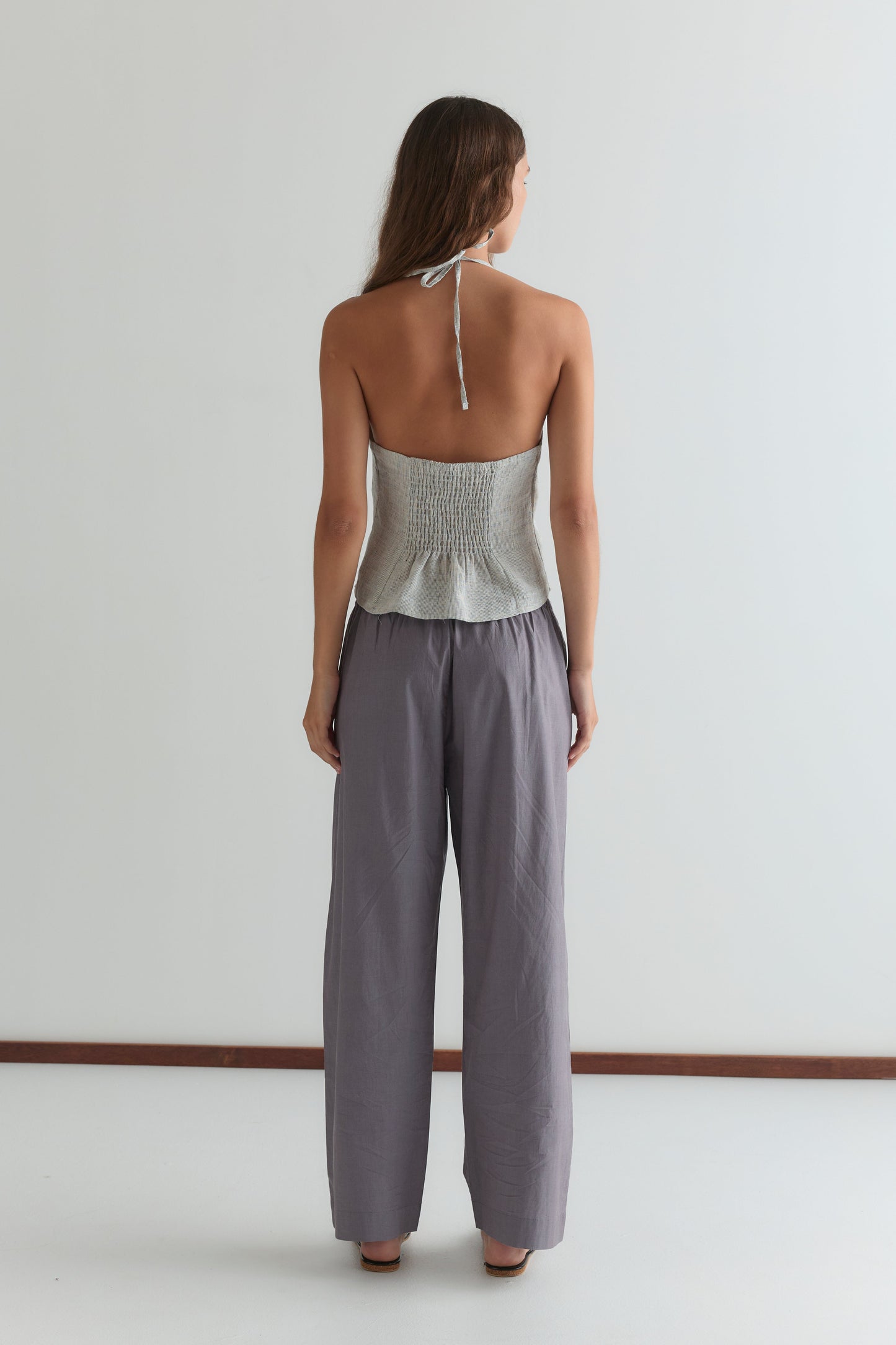 A woman stands facing away from the camera in a light gray halter top and Deiji Studios' Ease Trouser in Currant, shown in a minimal, light-filled room with a white wall and brown floor trim.